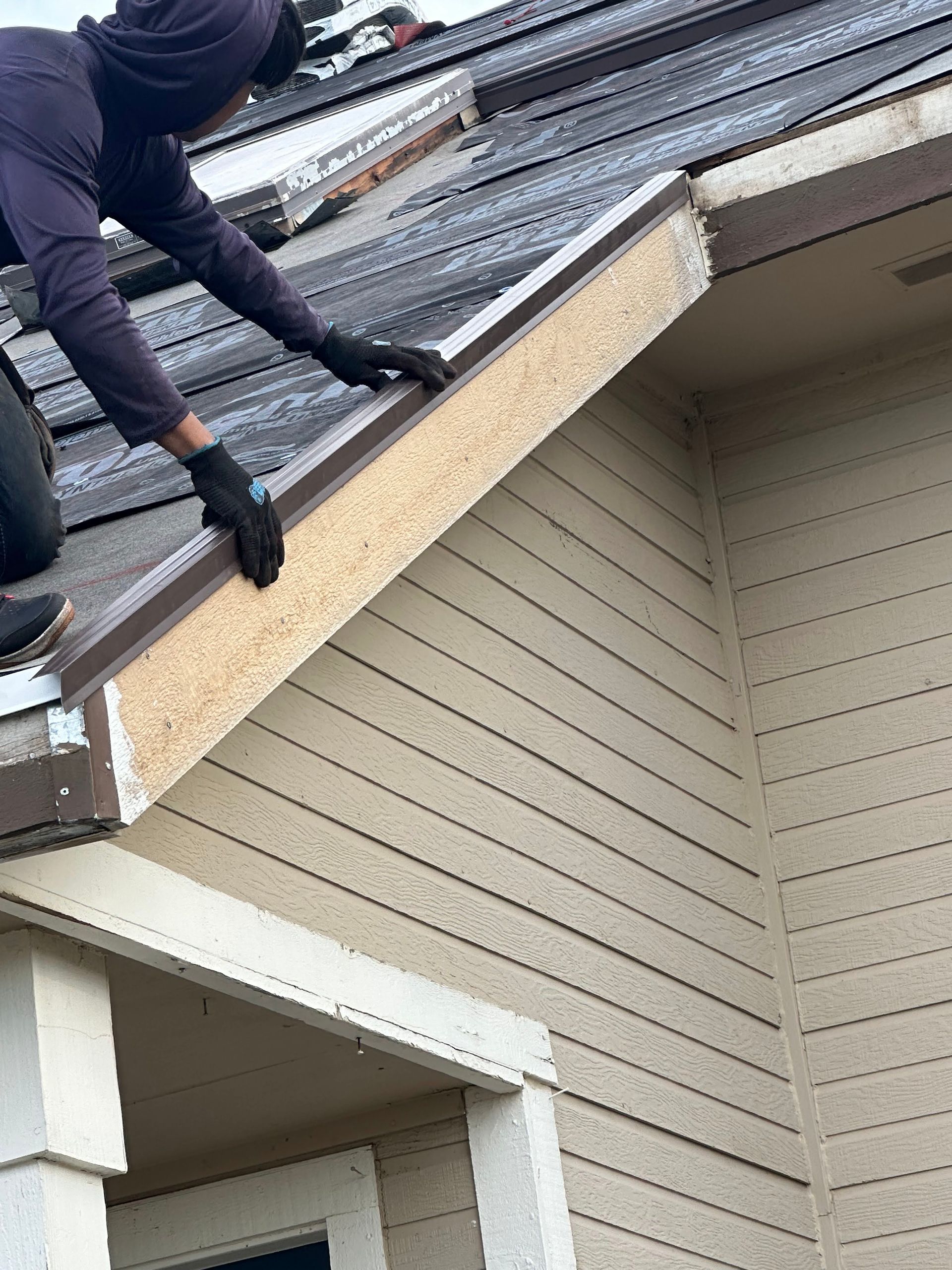 Worker on a roof installing shingles above a beige house exterior.