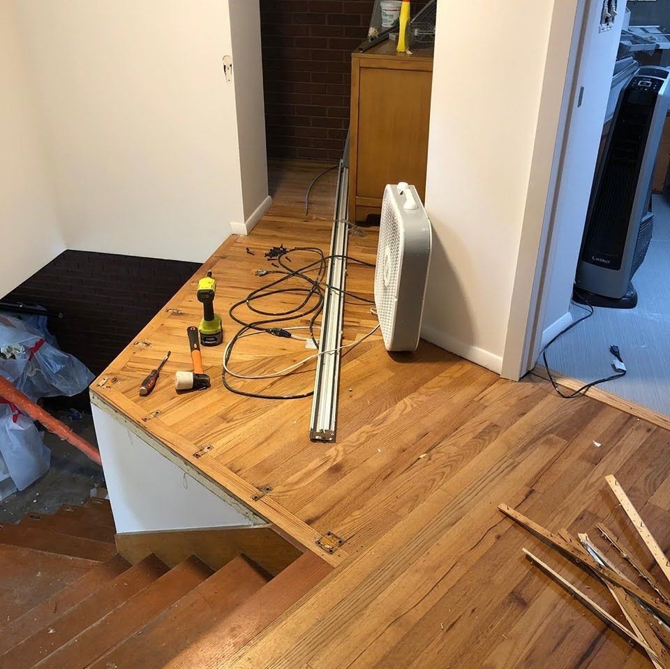 Wood floor renovation in progress with tools, cables, and a fan on unfinished subfloor.