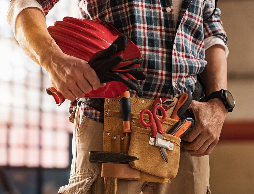 Carpenter holding red helmet and work gloves, wearing tool belt and plaid shirt.