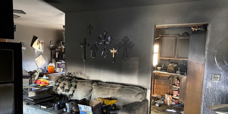 Interior view of a smoke-damaged home, showing living room and kitchen entrance. The walls are blackened.