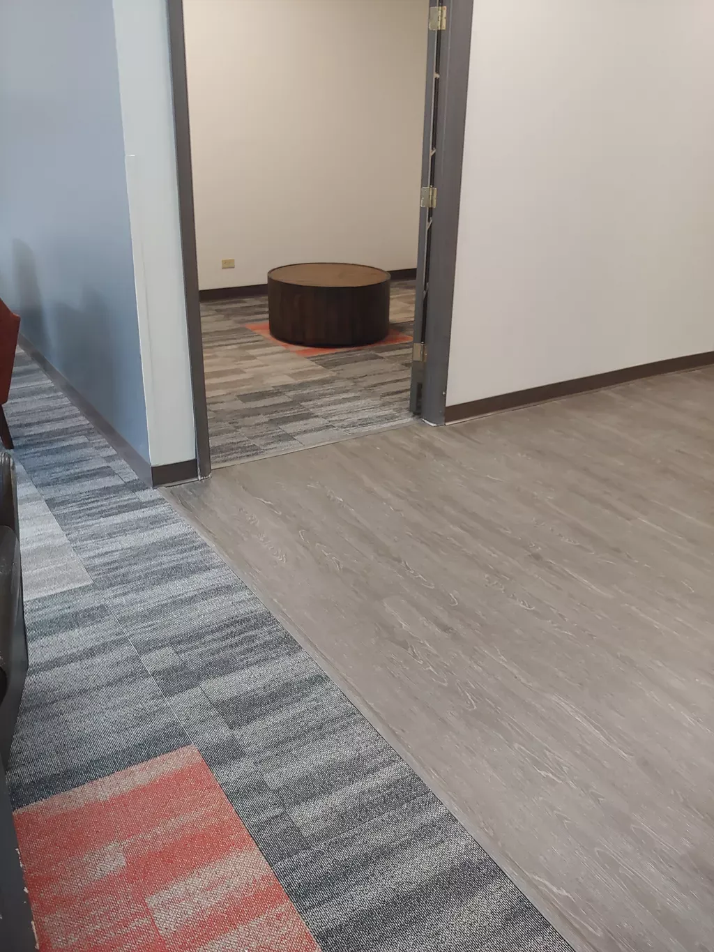 Doorway with gray wood-look floor leading to a room with patterned carpet and a brown circular ottoman.