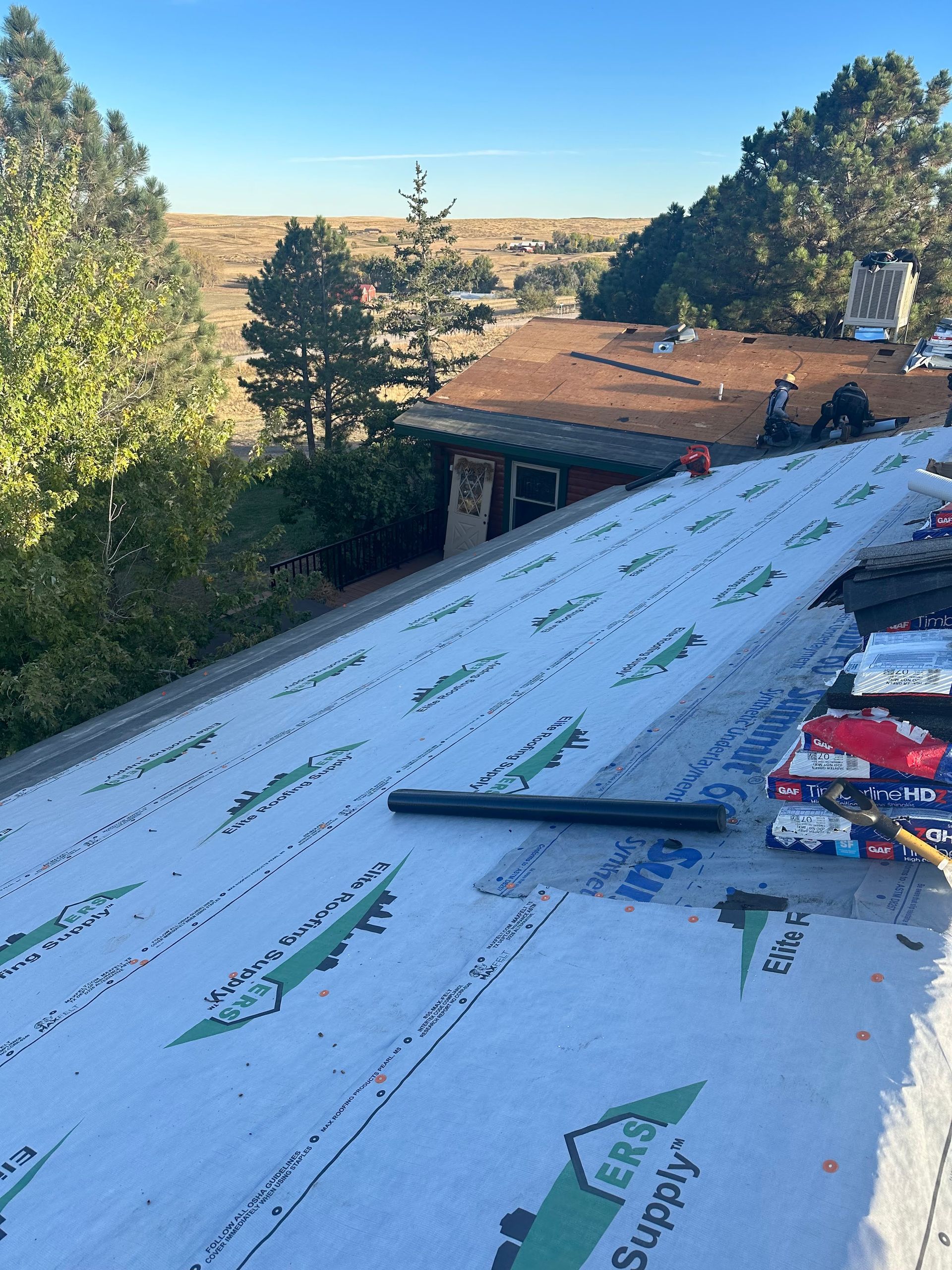 Workers installing white roofing underlayment on a sloped roof with tools, trees and a house in the background