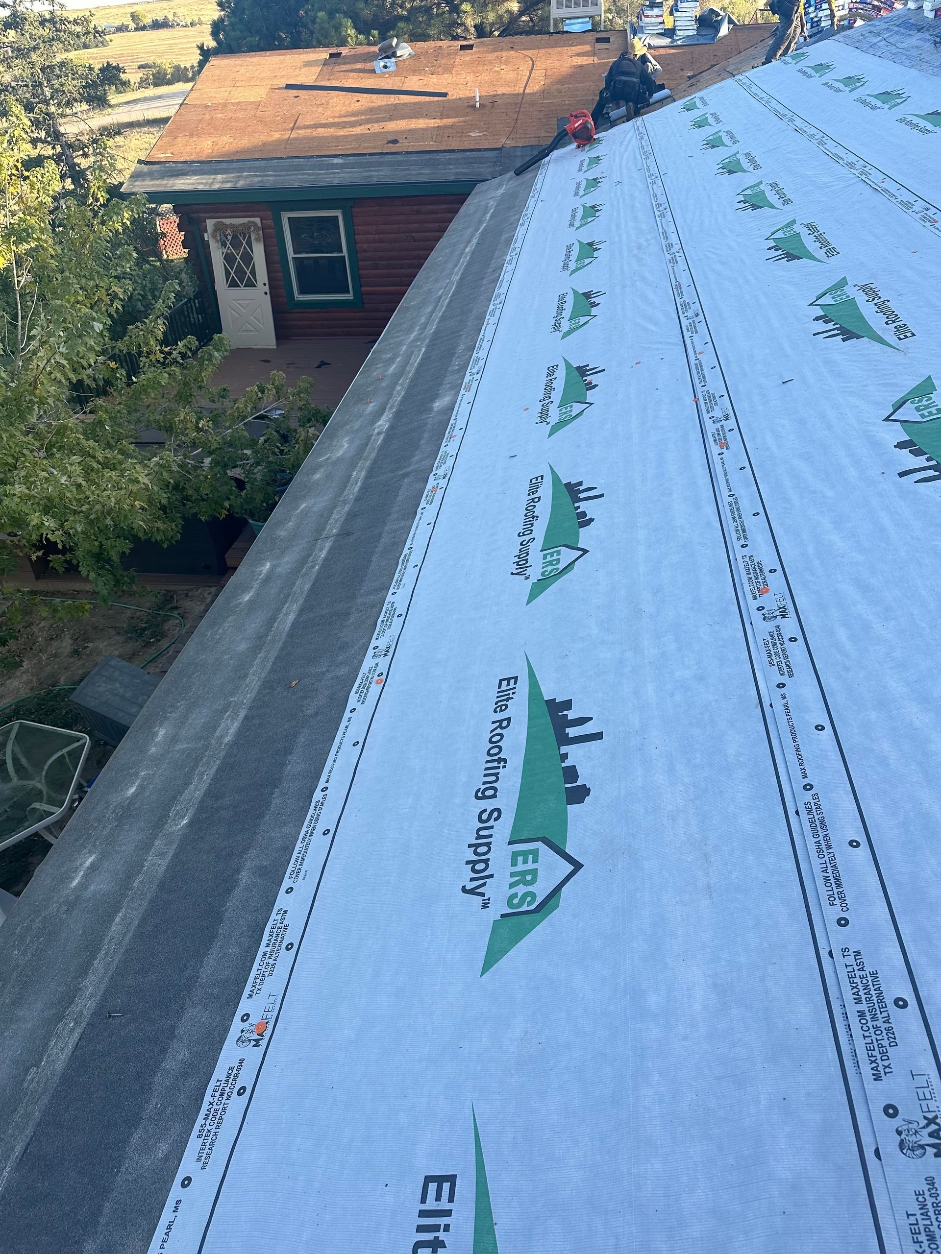 Roof under repair with white underlayment and green shingle pattern beside an older house.