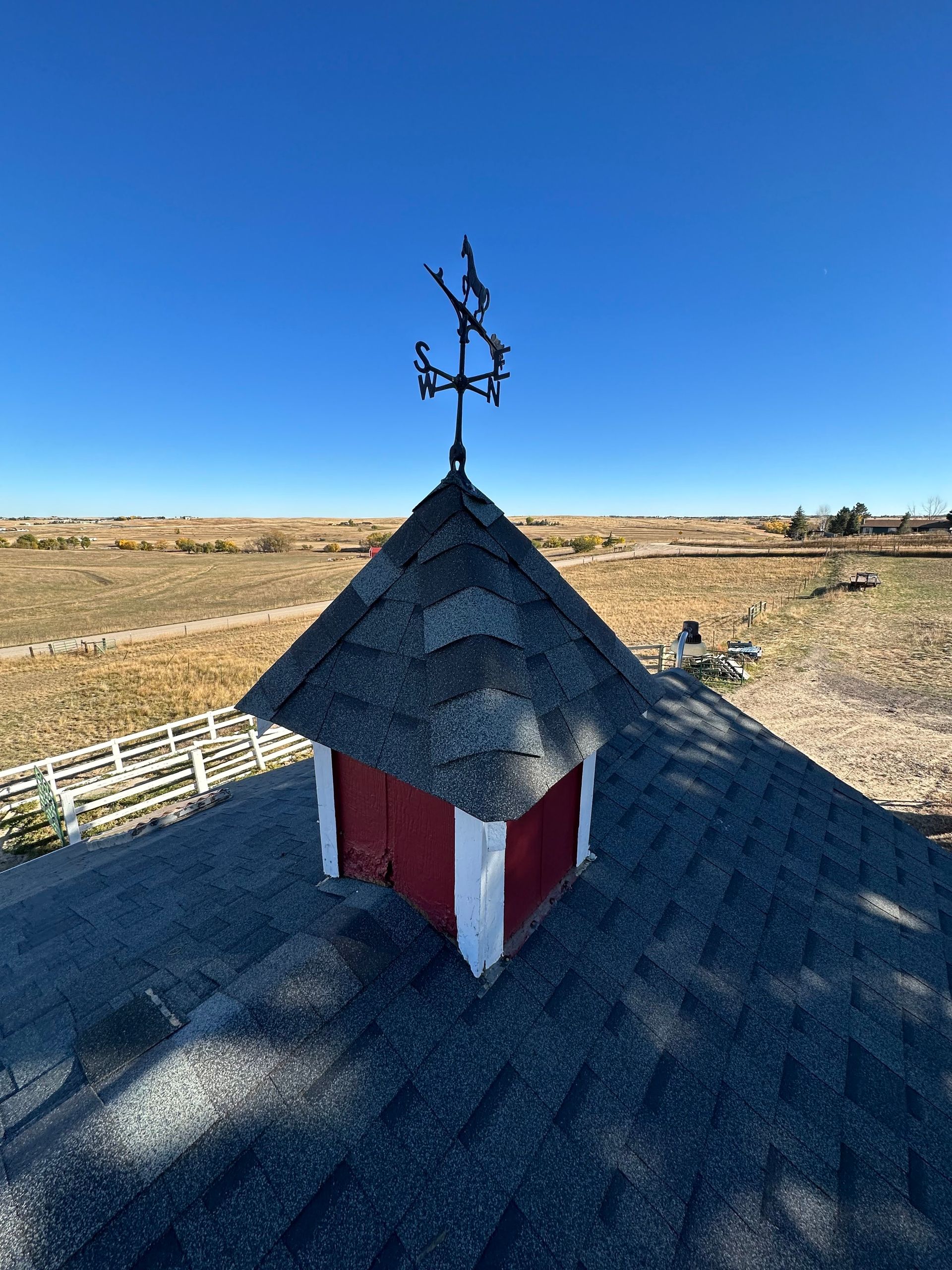 Small red and white cupola with dark shingled roof and weather vane on a rural rooftop under a clear blue sky