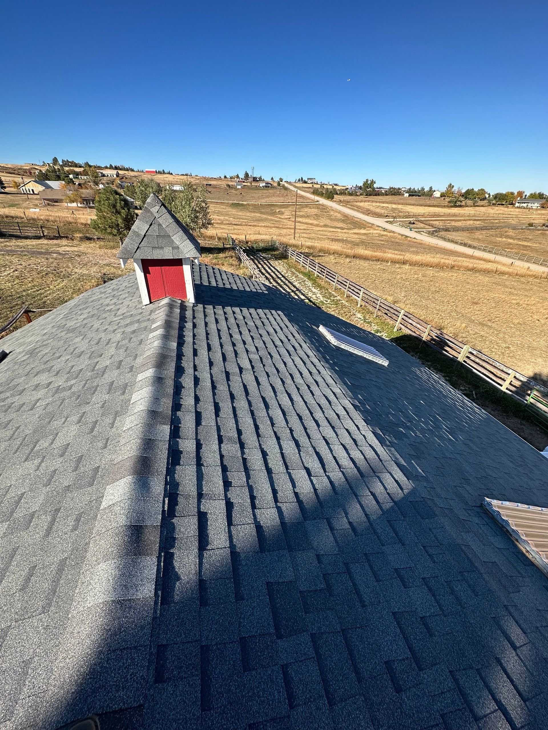 Roof with gray shingles and small red dormer under a clear blue sky, overlooking dry fields