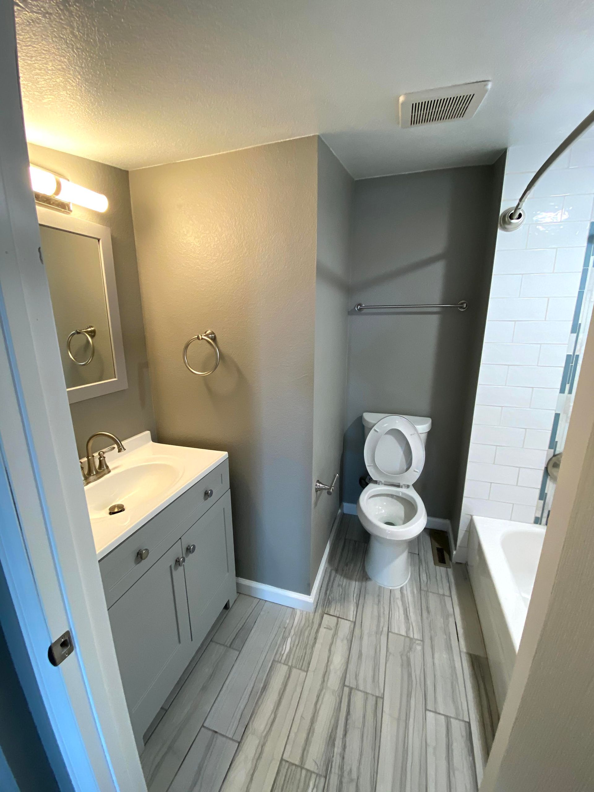 Small bathroom with vanity, mirror, toilet, tub, and gray tile flooring.