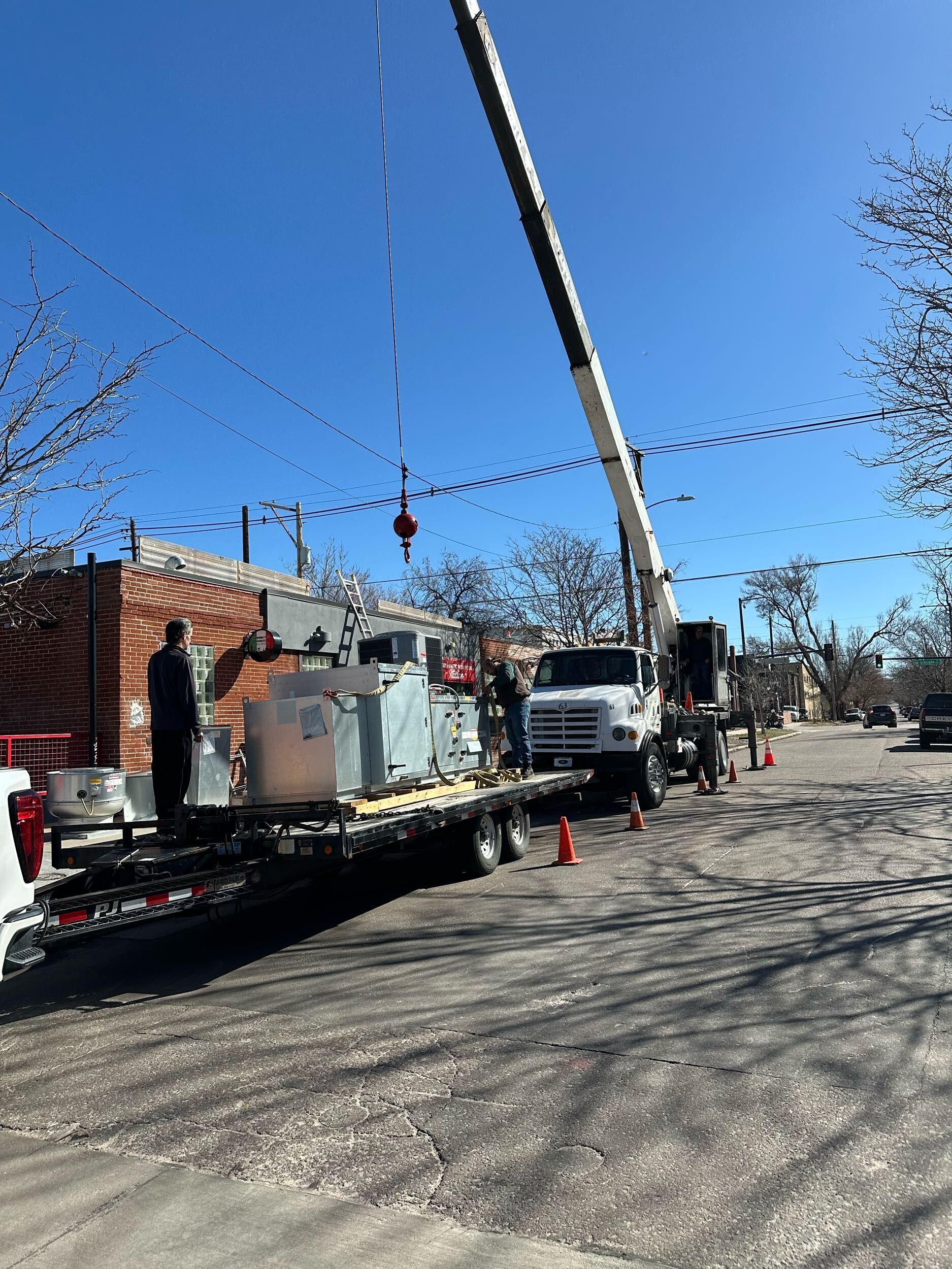 Crane lifting equipment onto a flatbed truck on a city street under a blue sky