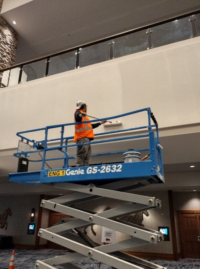 Worker on blue scissor lift inside a building, reaching up to inspect the ceiling.