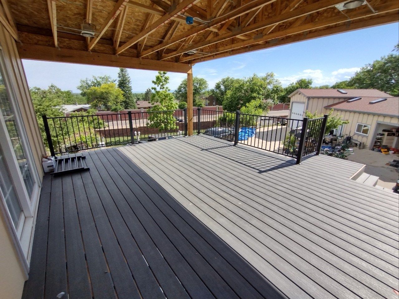 Covered wooden deck with gray flooring, railing, and view of houses and trees below