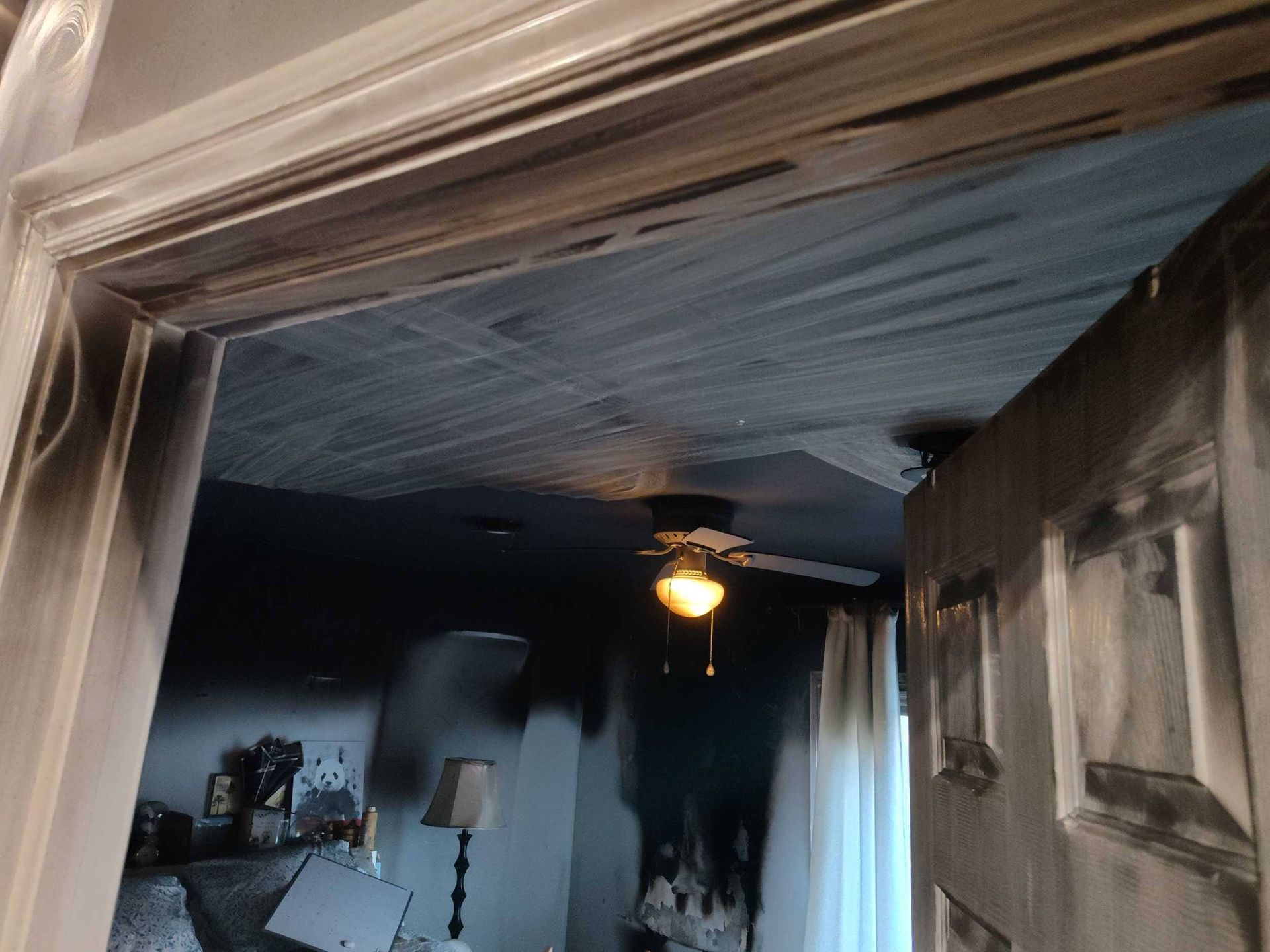 Dramatic dark bedroom with a ceiling fan light, white curtains, and charred-looking walls and doorway