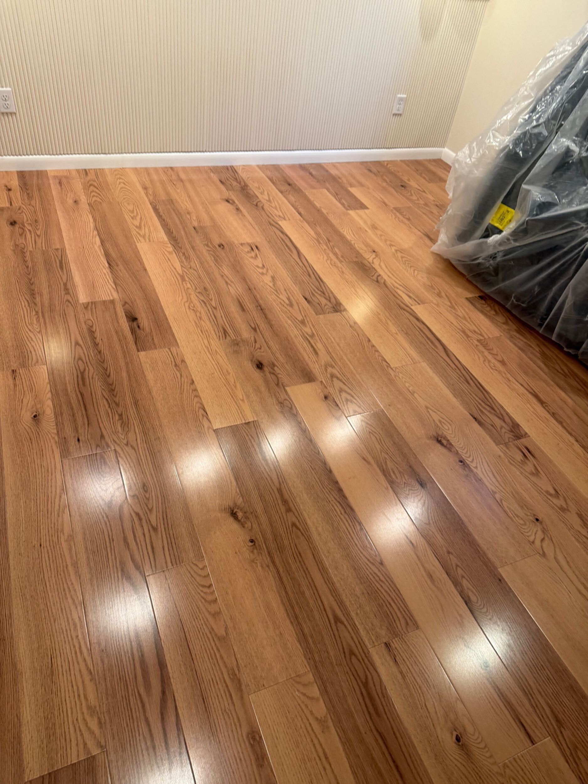 Empty room with glossy hardwood floor and white textured walls, with a wrapped item leaning in the corner.