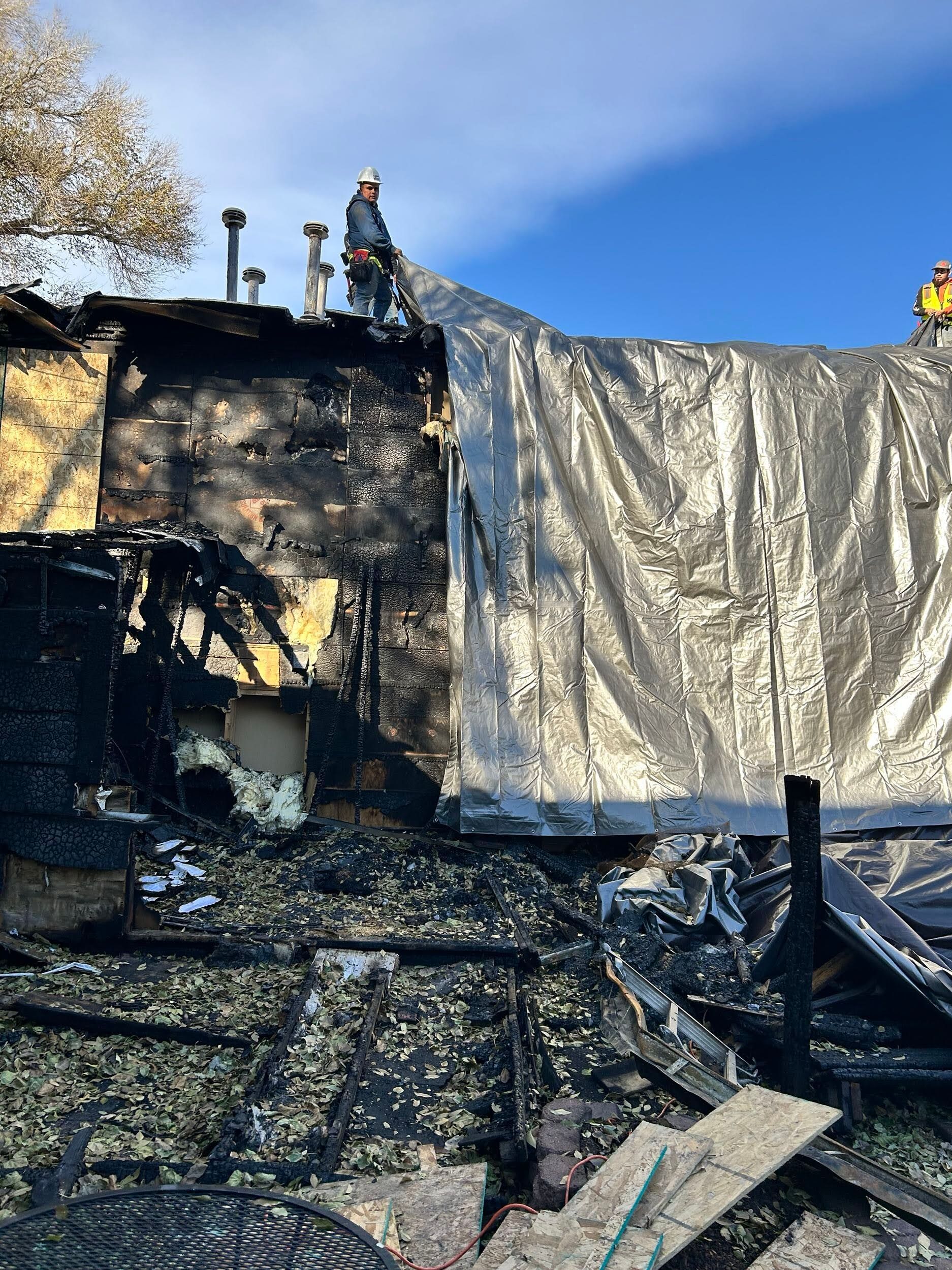 Burned building with collapsed roof and debris, firefighters standing on top under a blue sky