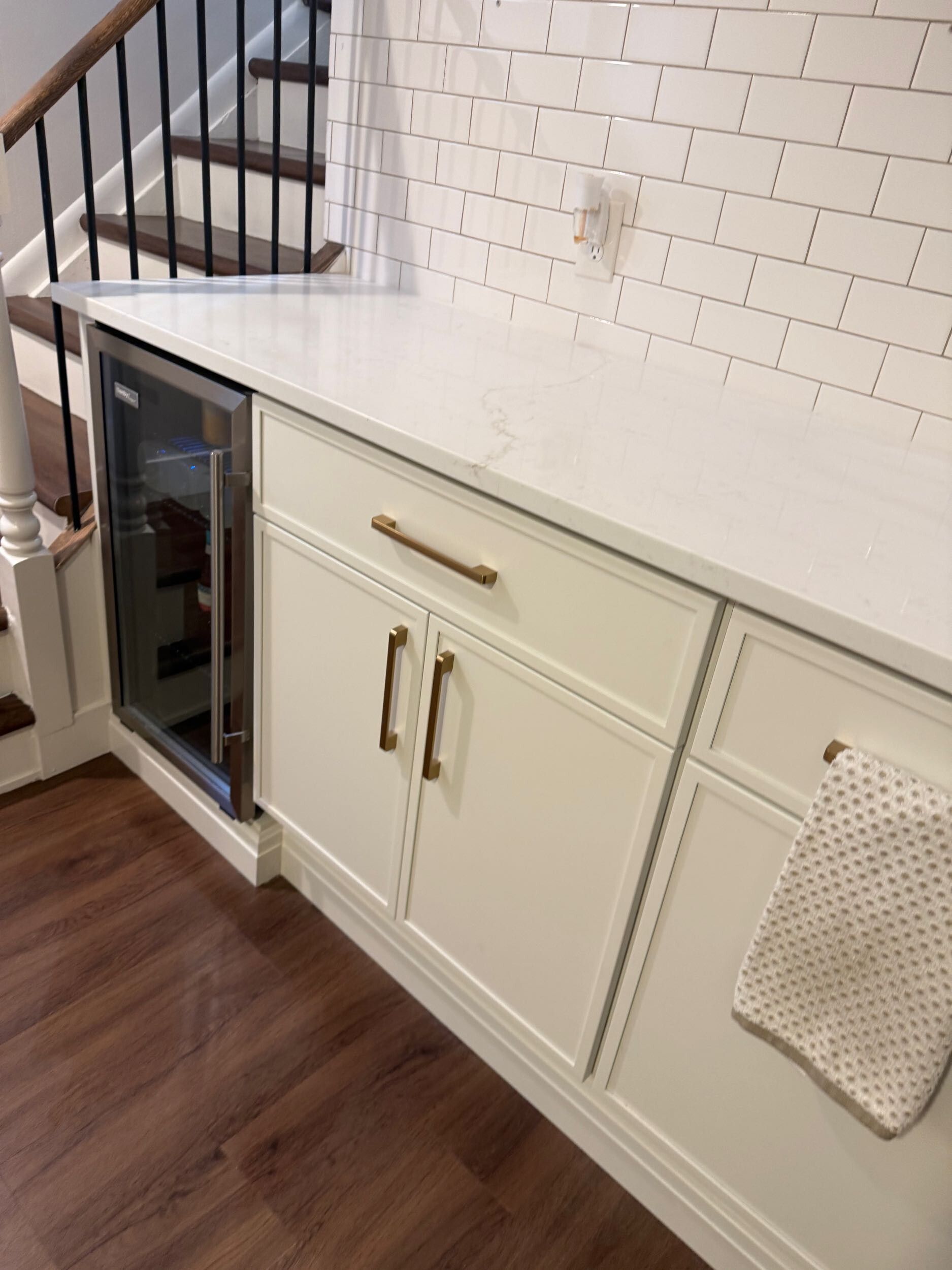 Built-in cream cabinet with small wine fridge under stairs, against white brick wall and dark wood floor