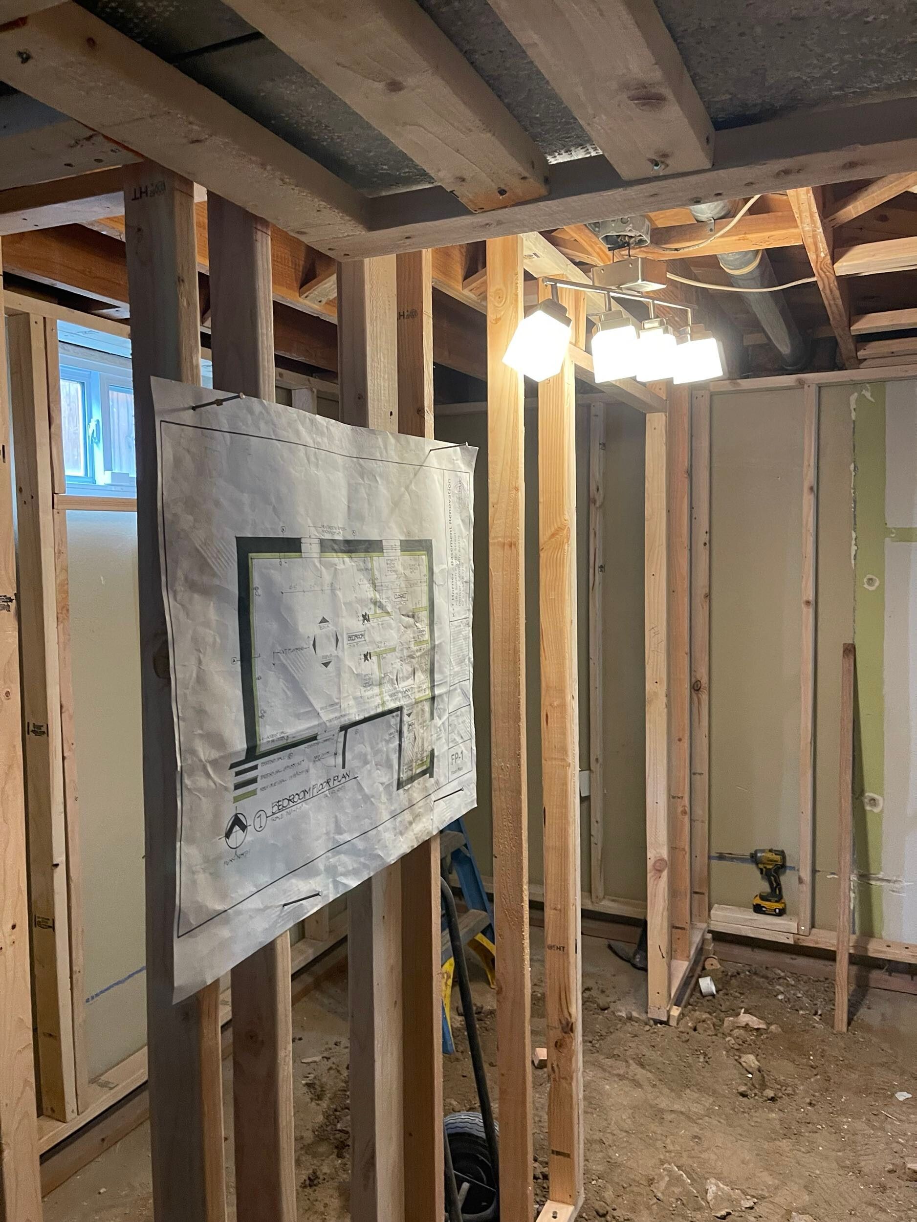 Interior of a stripped room with exposed studs, hanging light bulb, and a taped wall plan drawing.