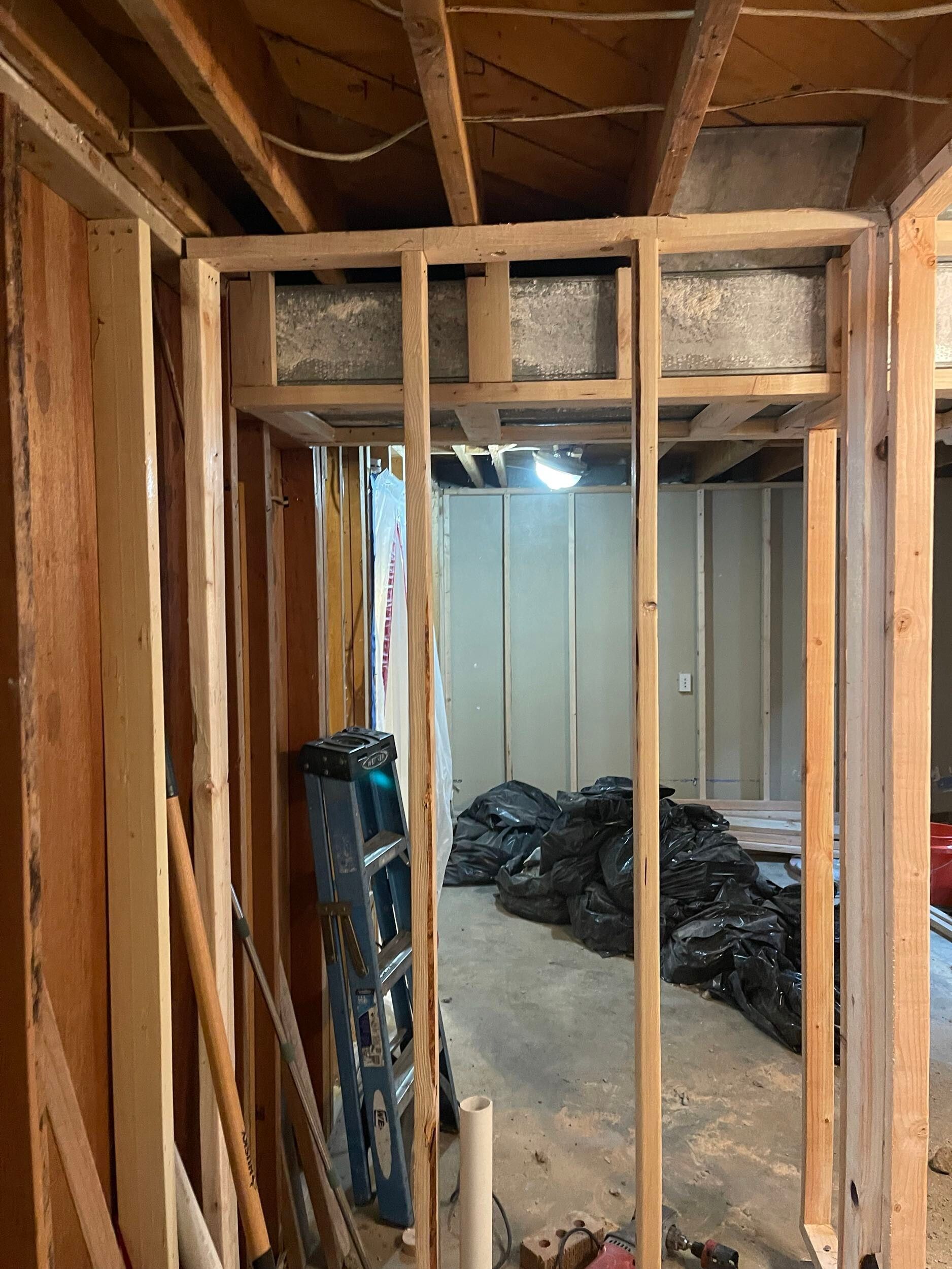 Unfinished basement framing with exposed wooden studs and ceiling beams, view into a room with debris bags.