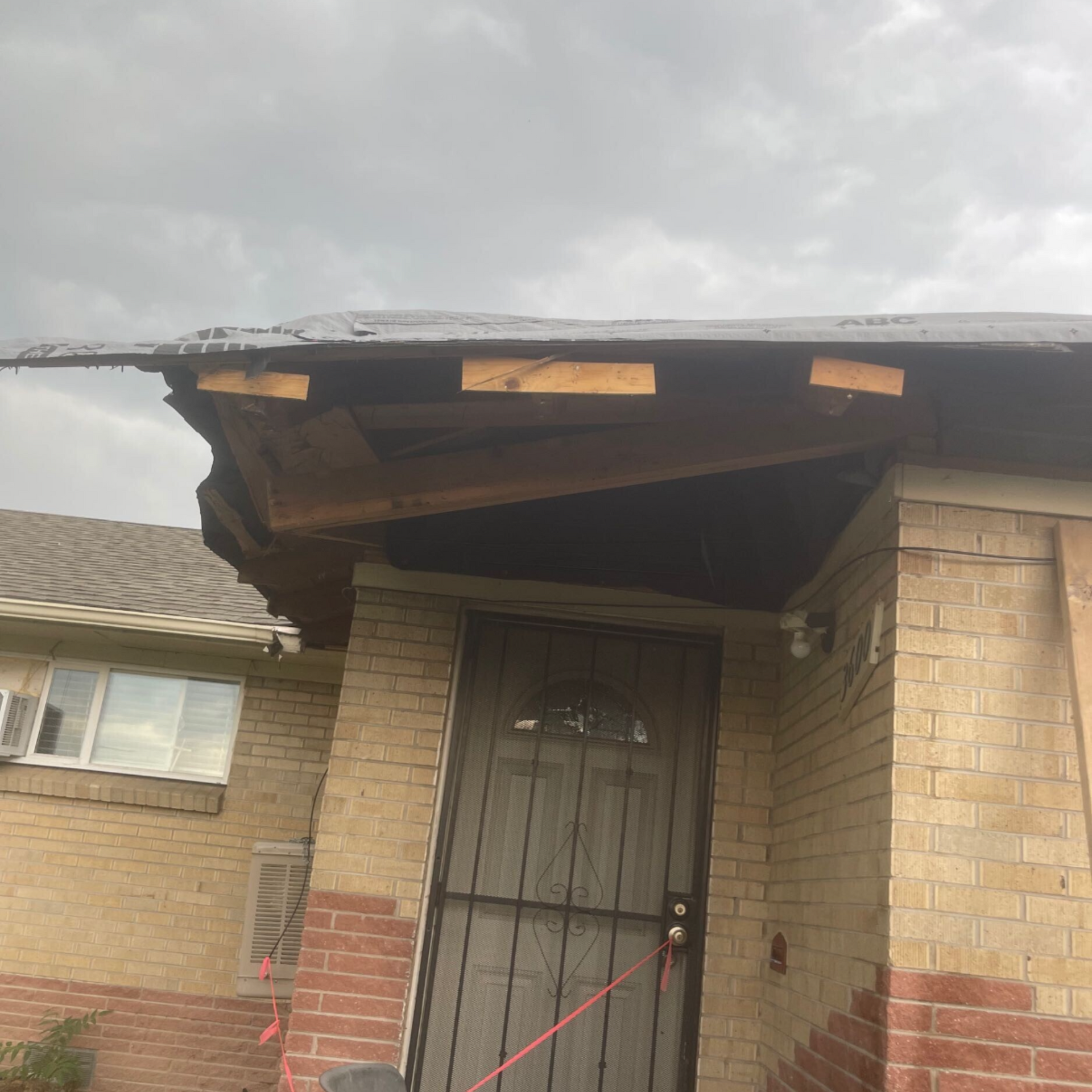 Damaged front porch roof with exposed wood above a brick house entrance and metal security door