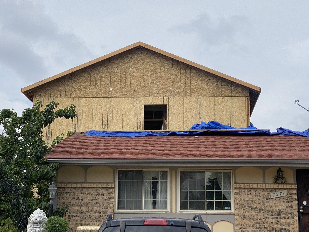 Two-story house with tan siding and a blue tarp on the roof under a cloudy sky