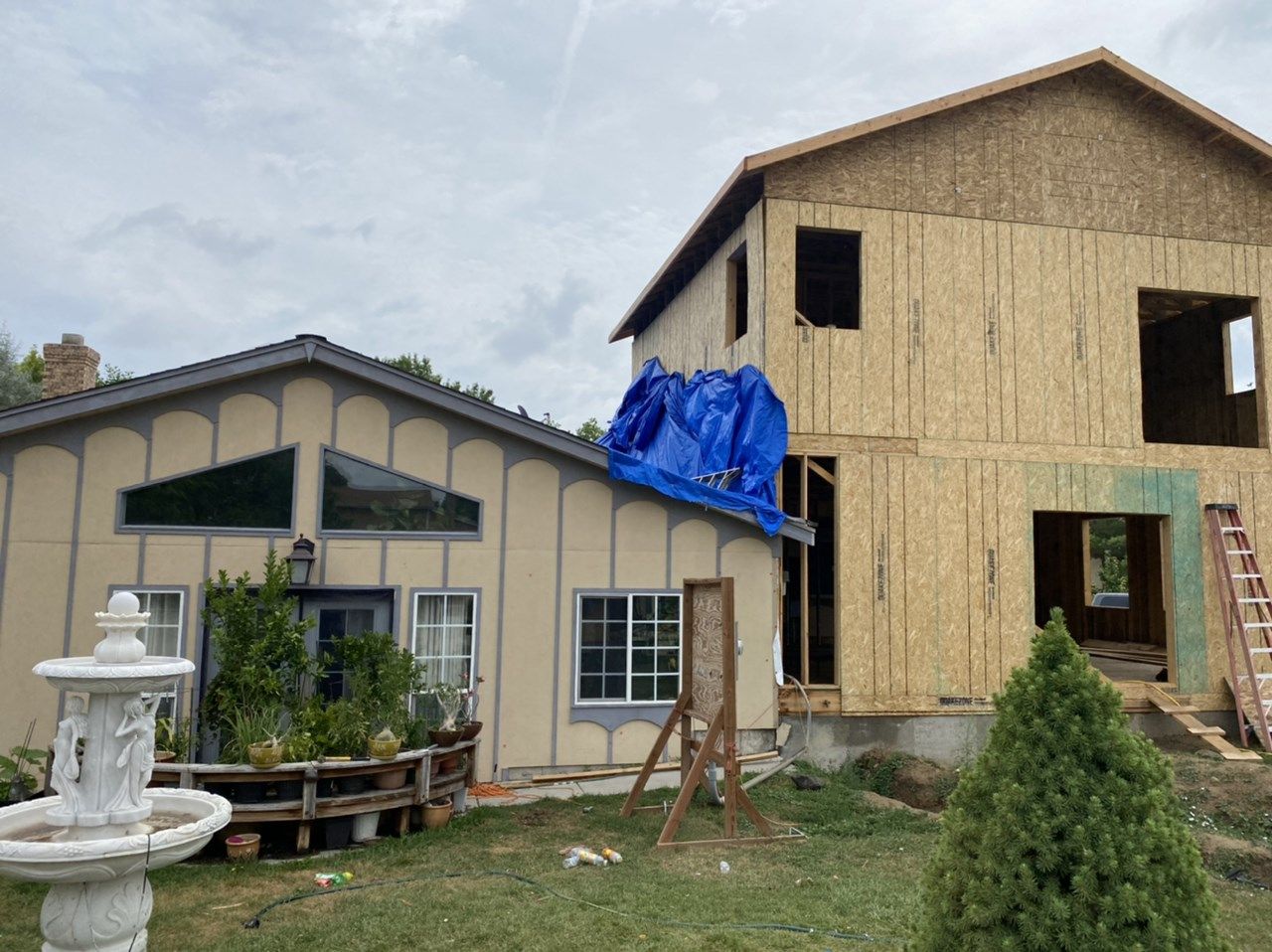 Two-story house under construction beside a finished beige house, with a blue tarp and wooden scaffolding.