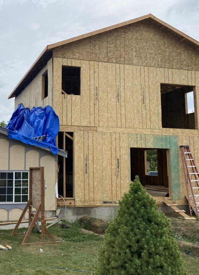 Two-story house under construction with plywood siding, blue tarp, and a ladder by the front opening.