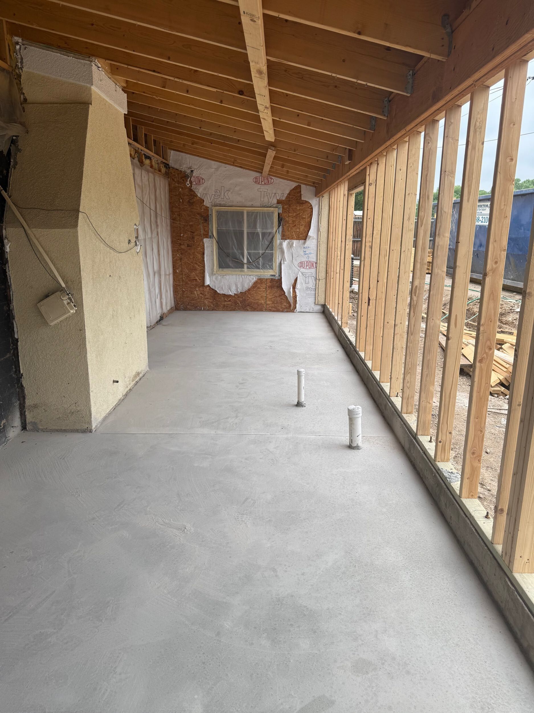Covered porch under construction with concrete floor, exposed wood framing, and window opening.