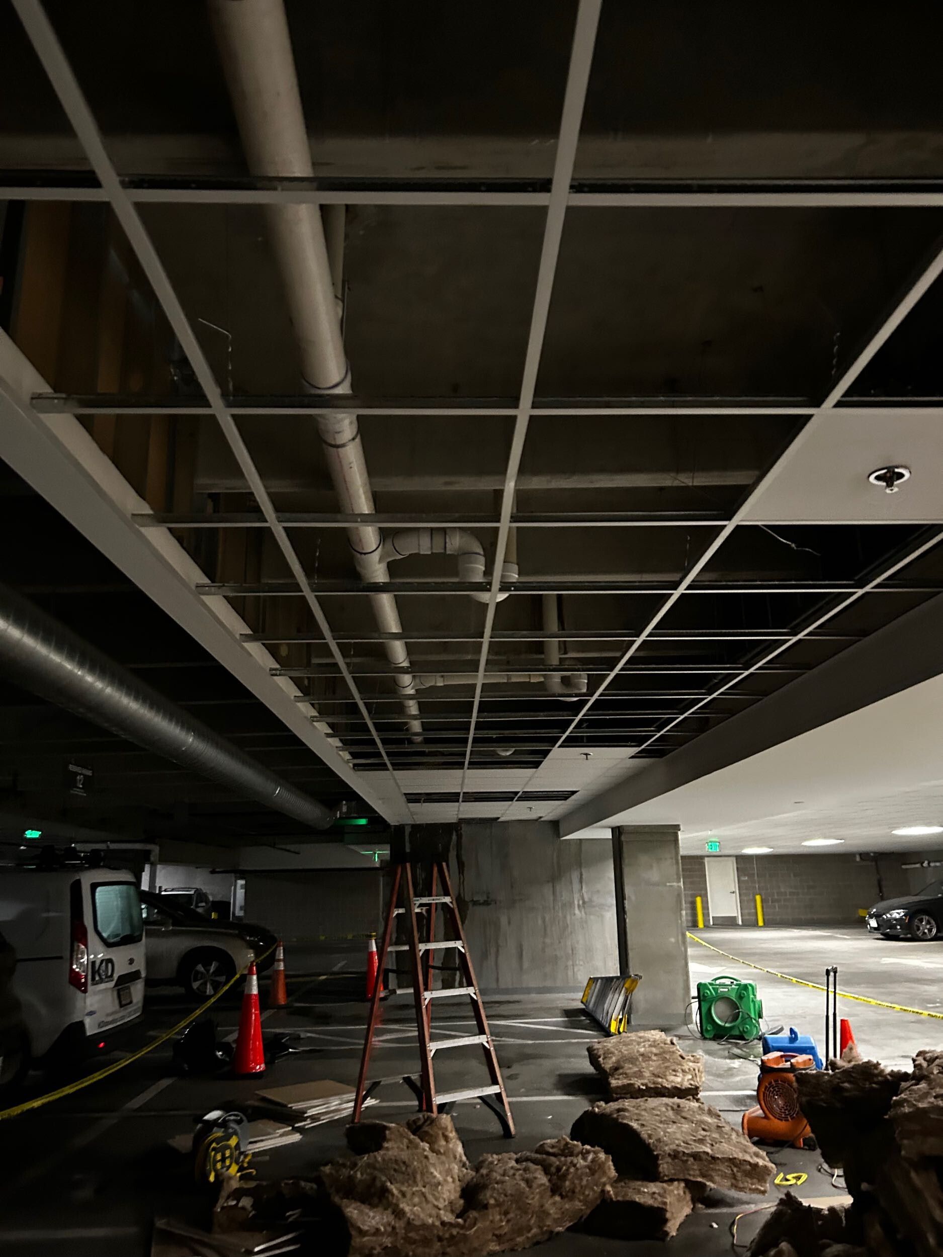 Underground parking garage under construction with exposed ceiling pipes, ladders, cones, and building materials