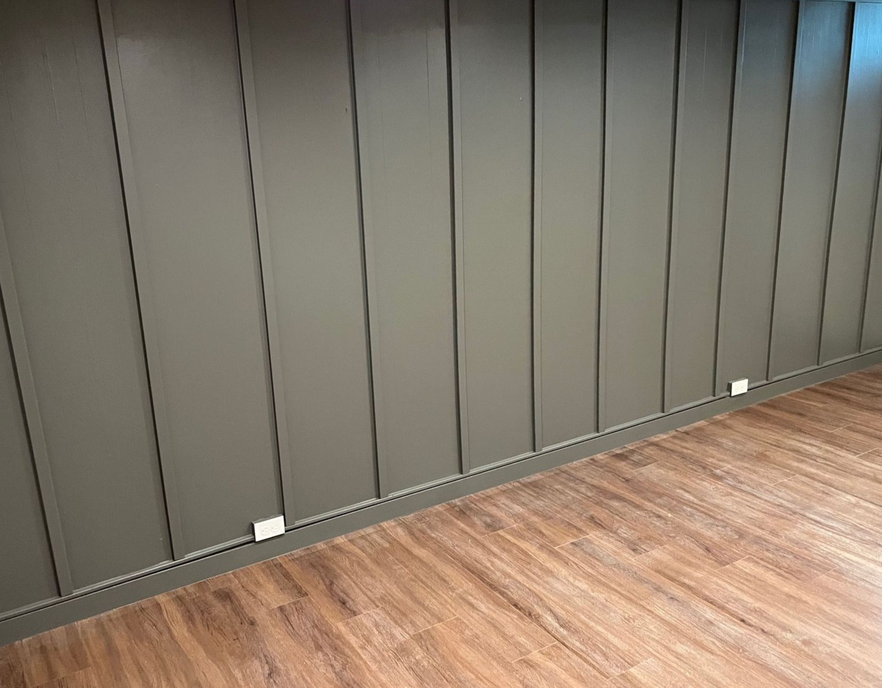 Dark paneled wall with wood flooring and small floor outlets along the base