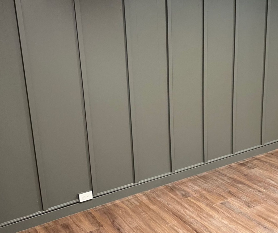 Gray paneled wall above wood flooring, with a small white outlet near the baseboard.