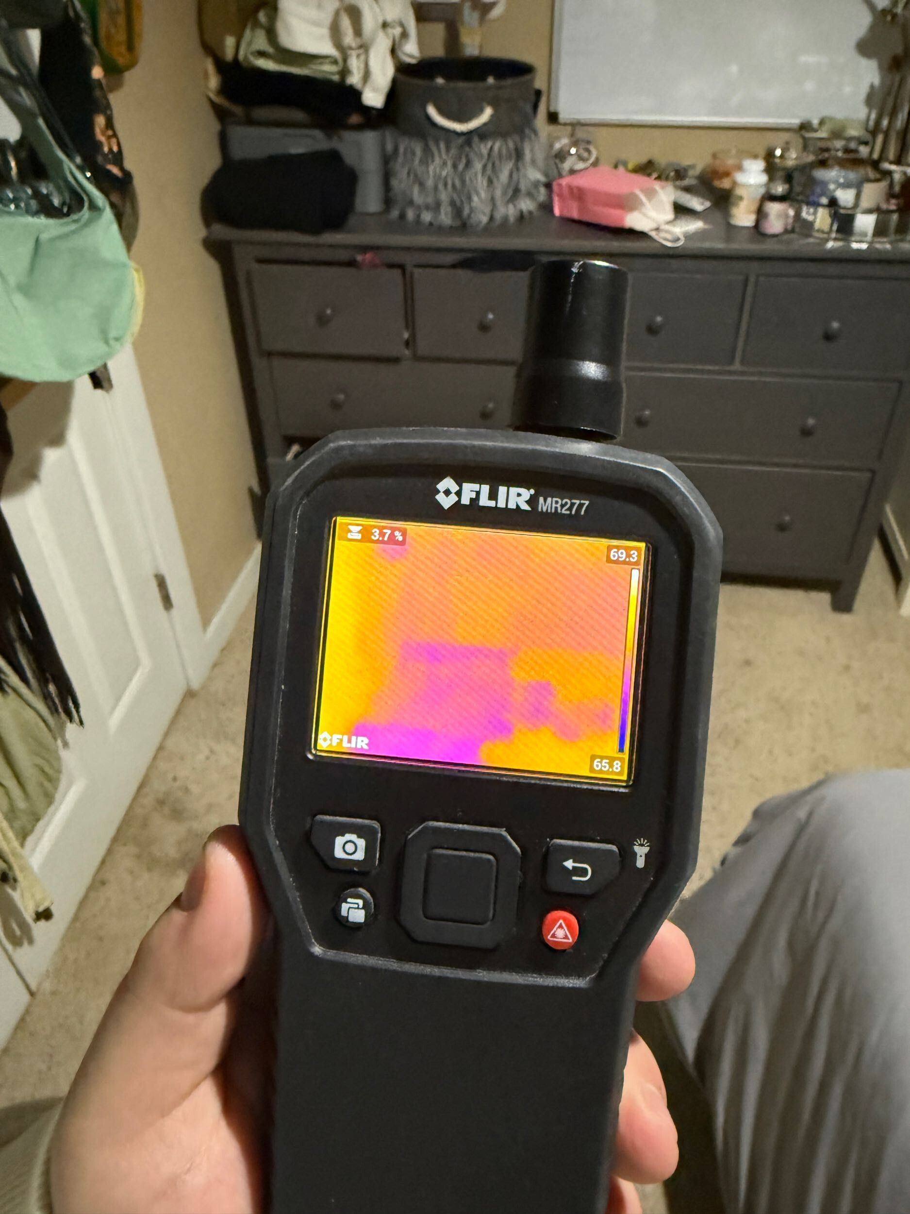 Hand holding a FLIR thermal camera in a bedroom, showing a pink-orange heat display.