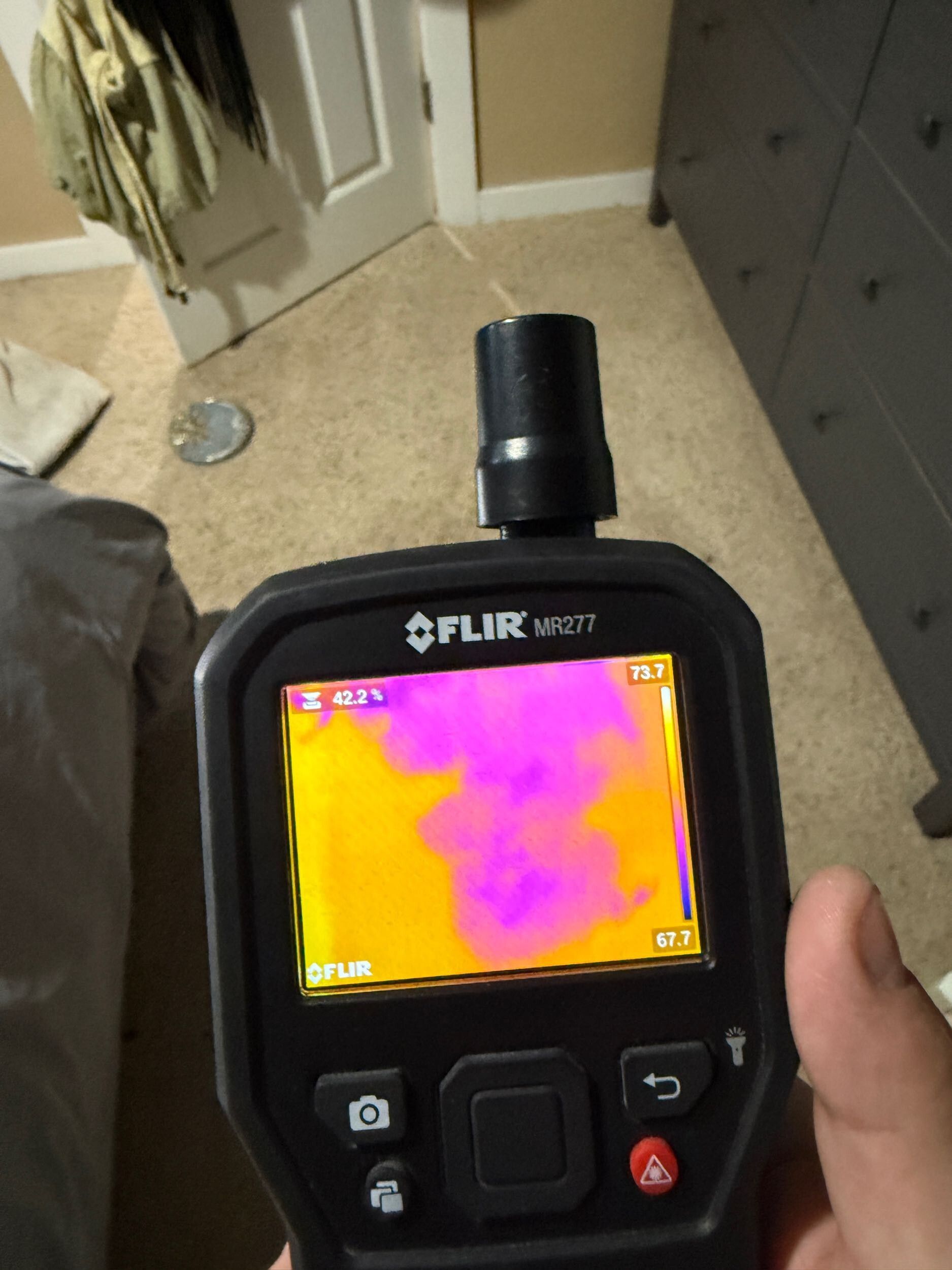 Handheld FLIR thermal camera showing a pink and orange heat map in a room with cabinets and a doorway.