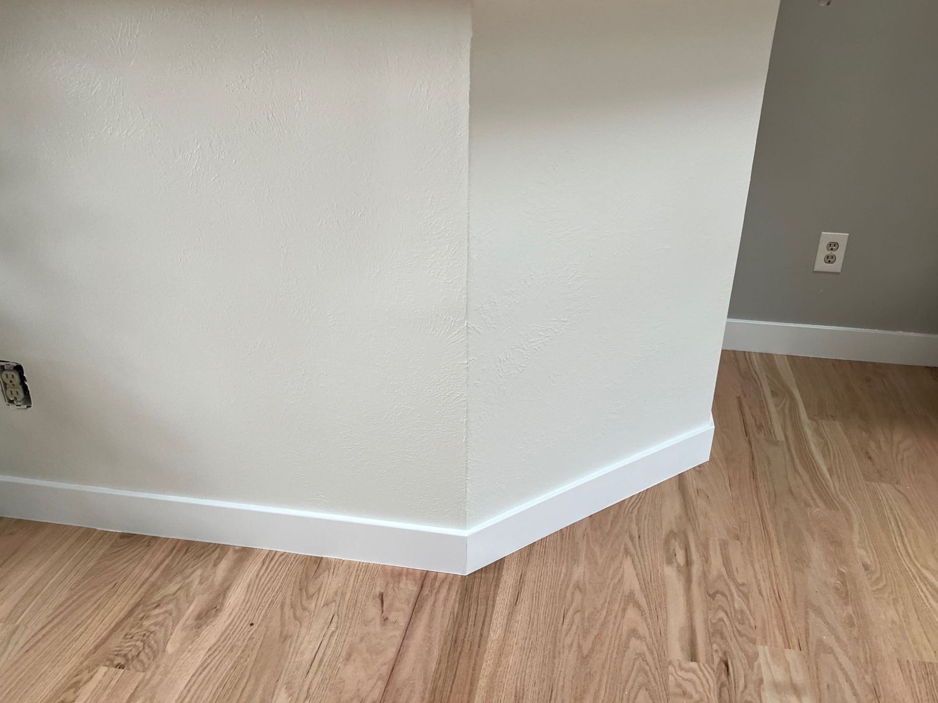 Empty room corner with white walls, baseboards, and light wood flooring.
