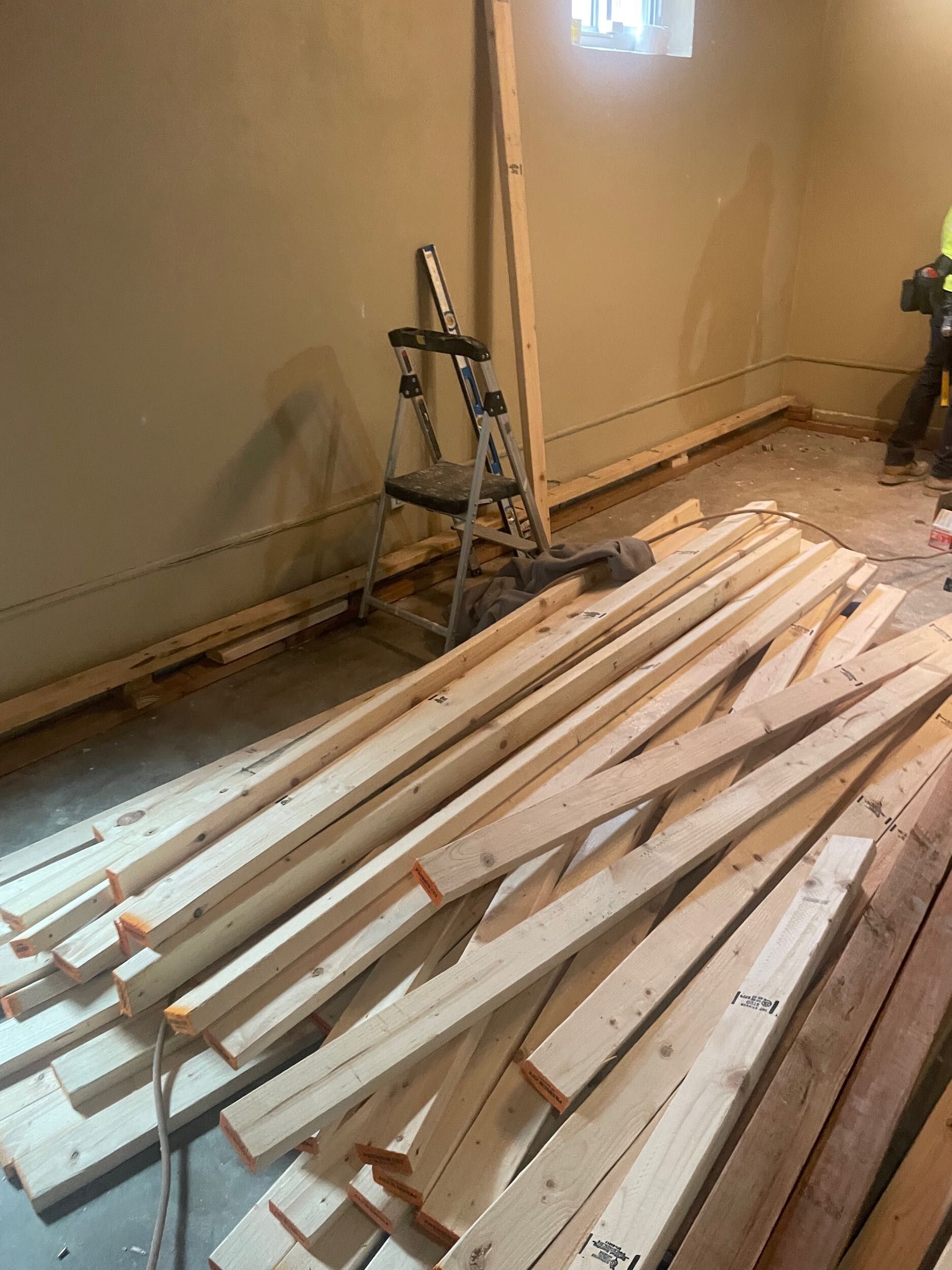 Long wooden boards stacked on a floor in a partially renovated room with a step ladder against the wall.