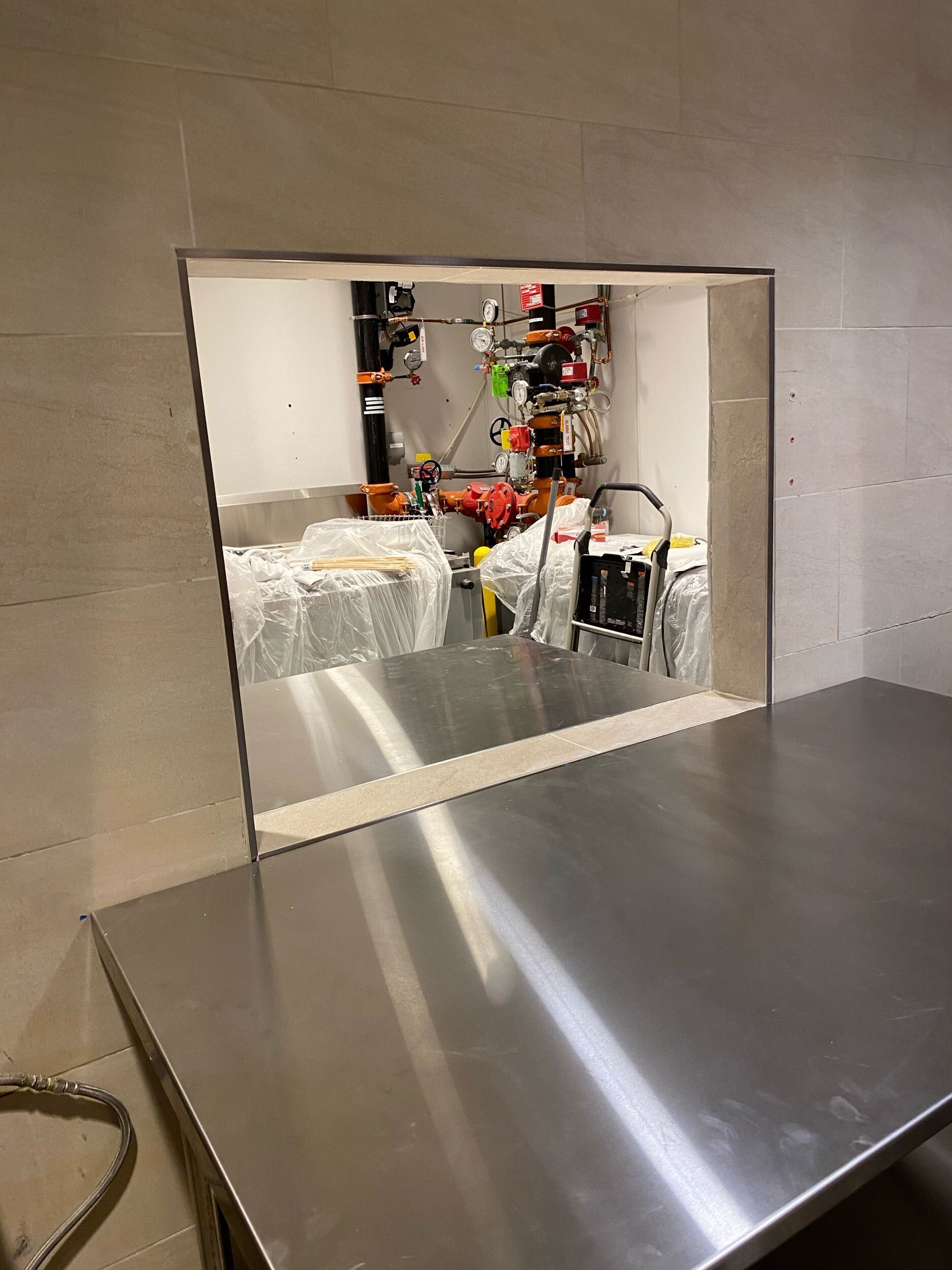 Stainless steel counter with a rectangular opening showing machinery and orange-clad workers behind it