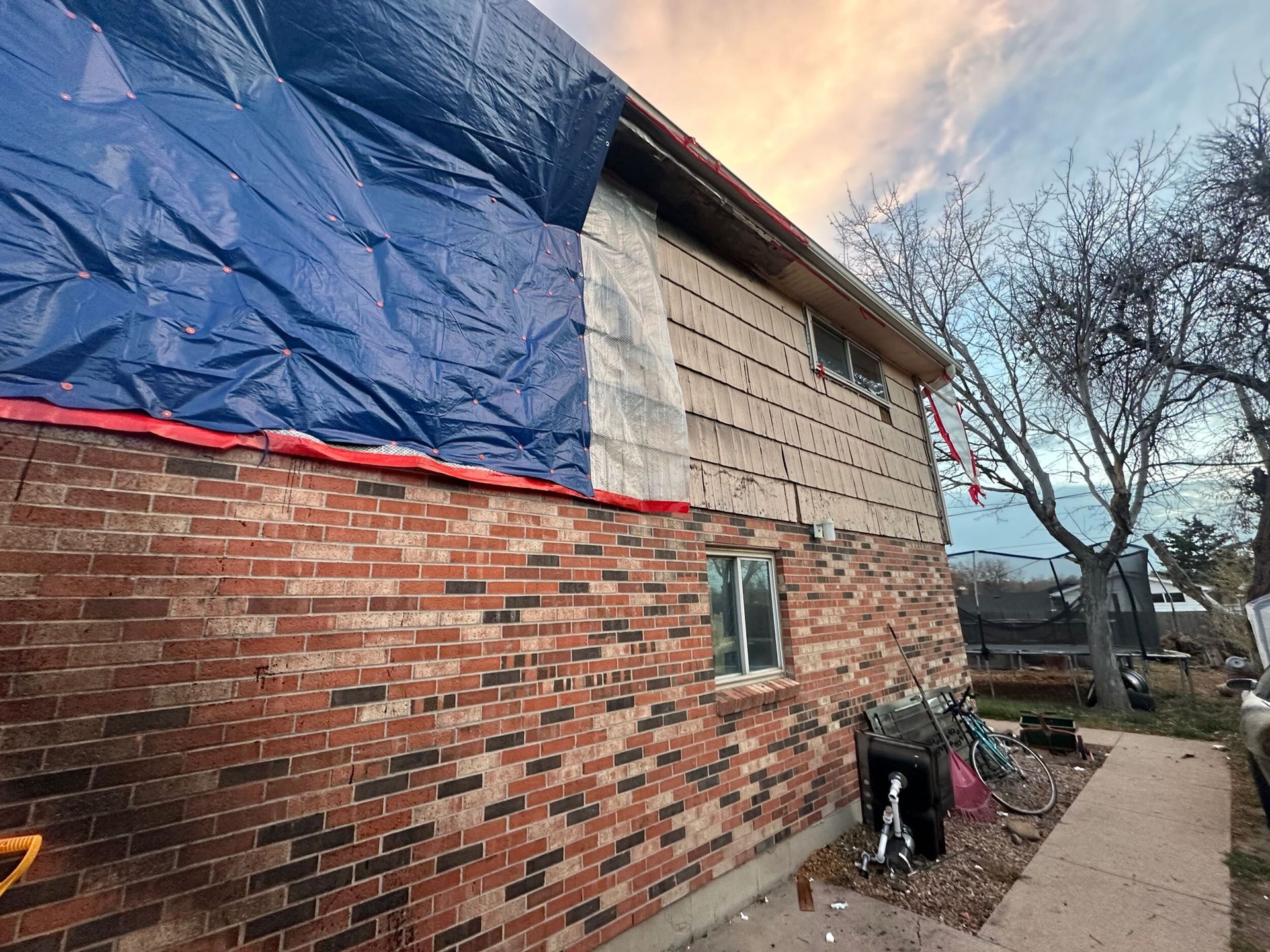 Brick house with blue tarp on roof, window, and a cluttered side yard beside a sidewalk