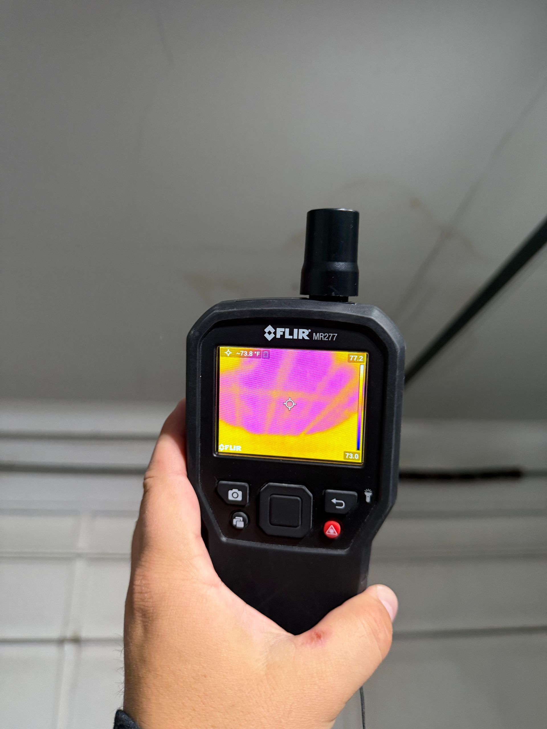 Hand holding a thermal camera showing a pink-and-yellow heat map on its screen in a gray room