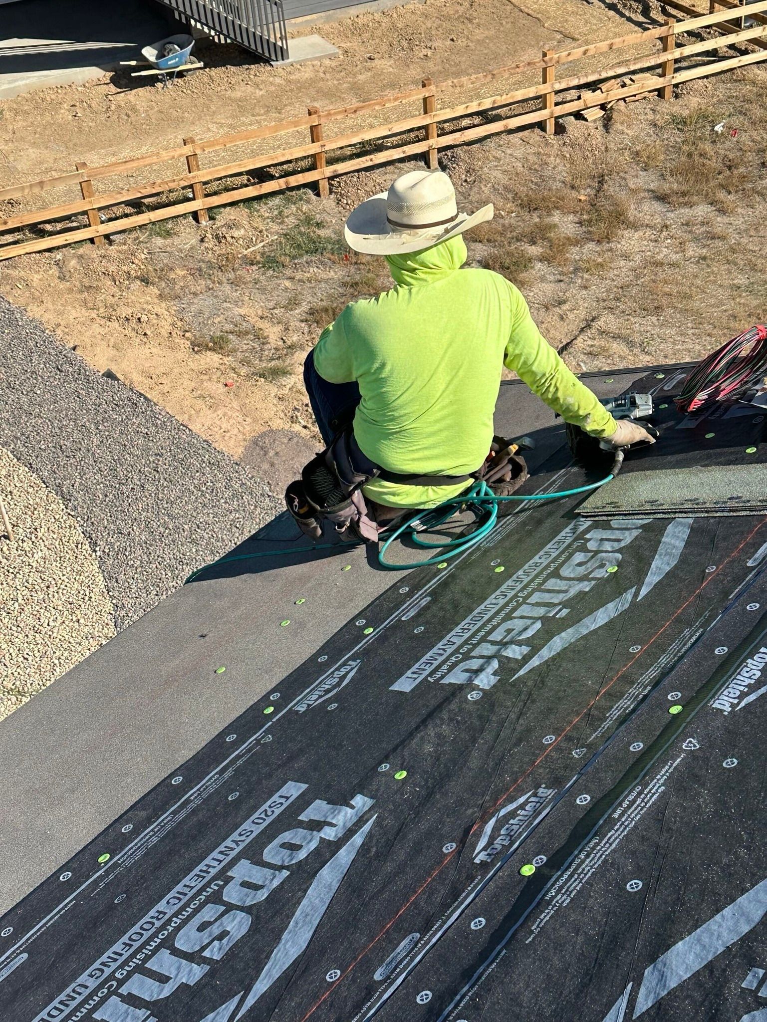Worker in neon green shirt and hard hat on a rooftop, handling ropes beside roofing materials.