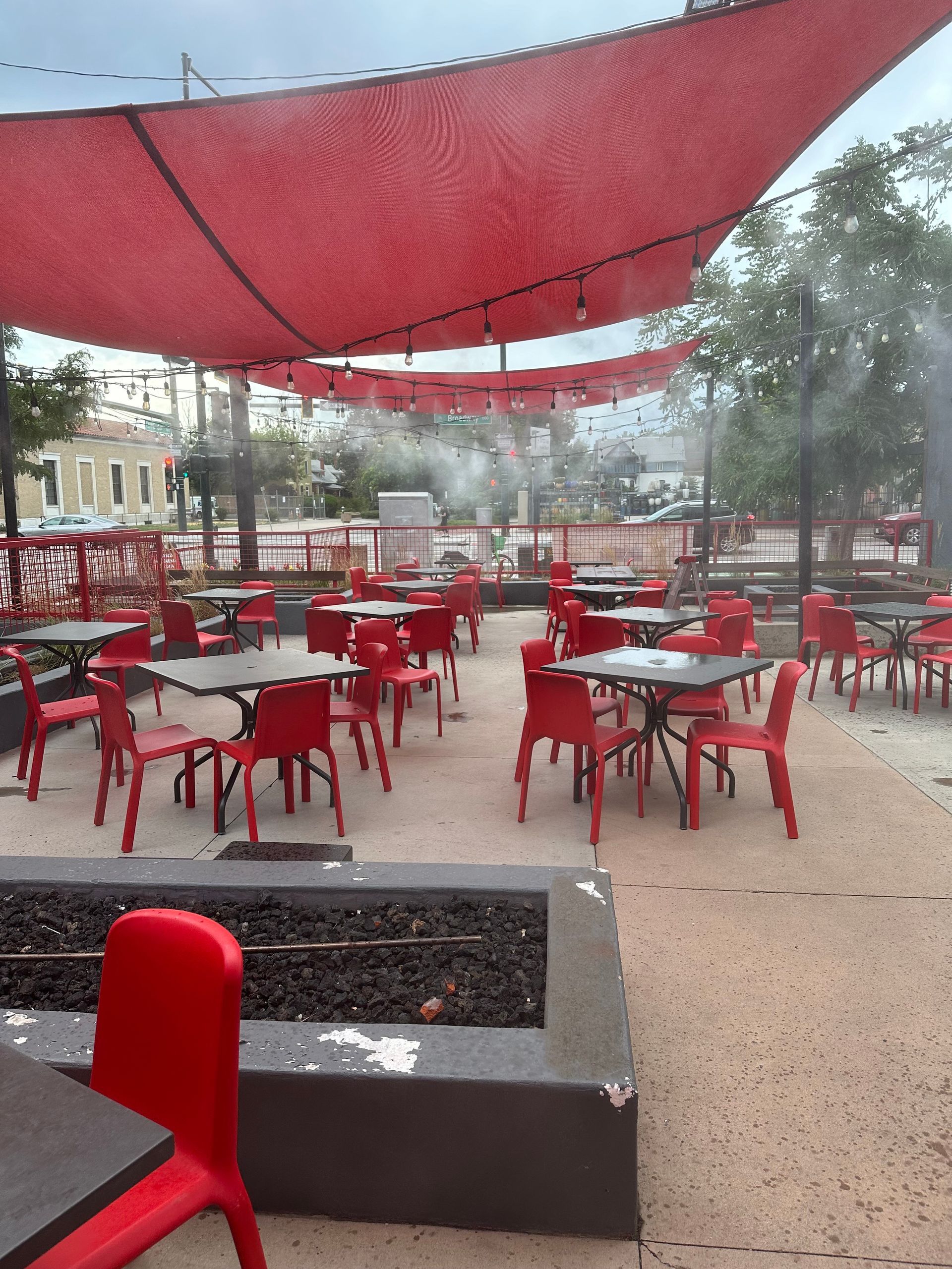 Outdoor café patio with red chairs and tables under red shade sails, seen in a rainy setting