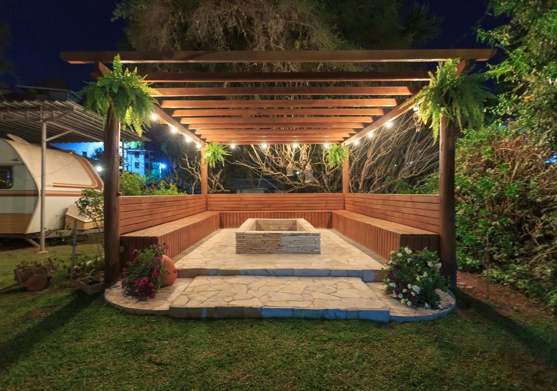 Nighttime outdoor patio with a wooden pergola, stone seating area, and surrounding greenery