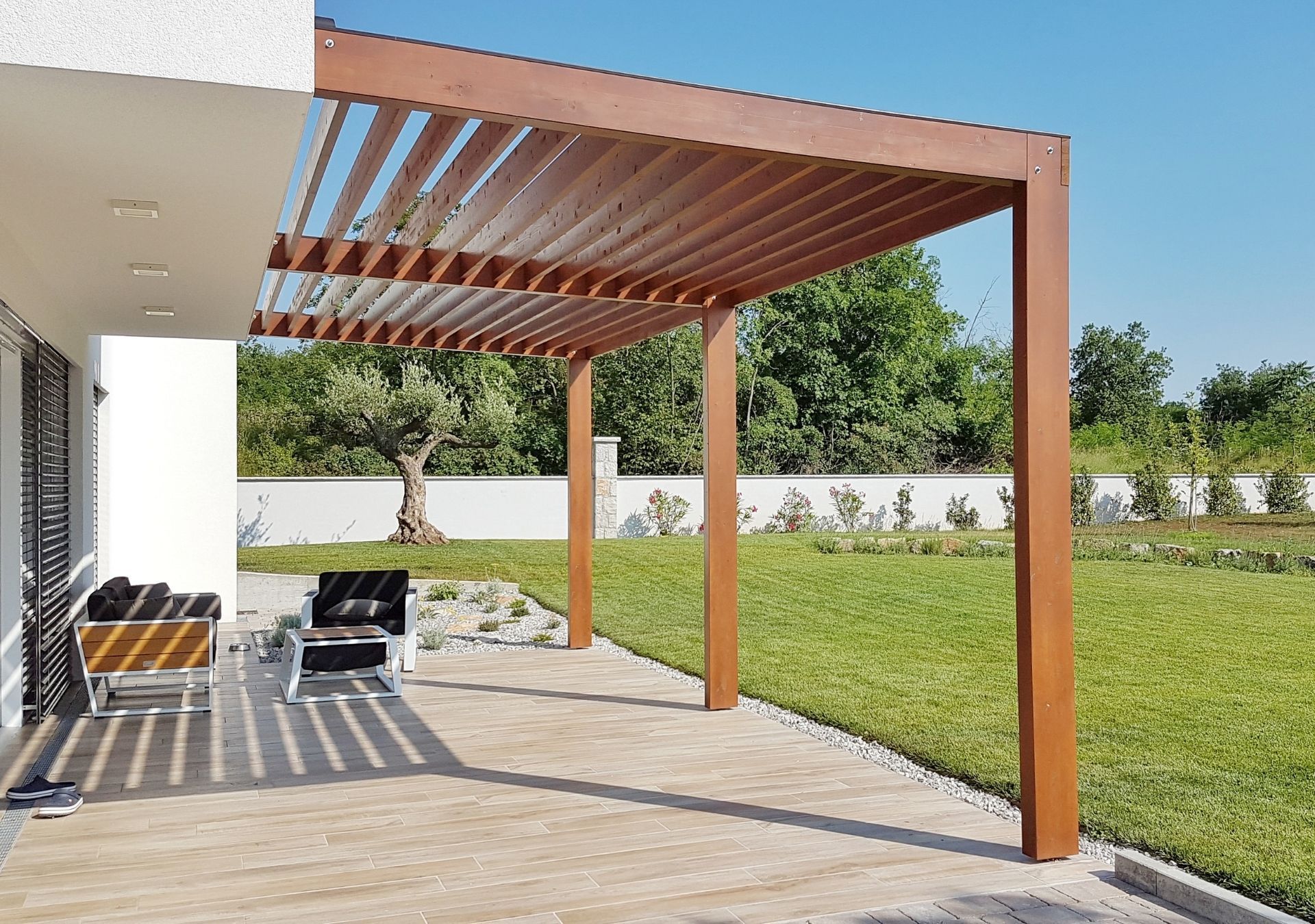 Wooden pergola over a patio with outdoor chairs beside a lawn and white wall