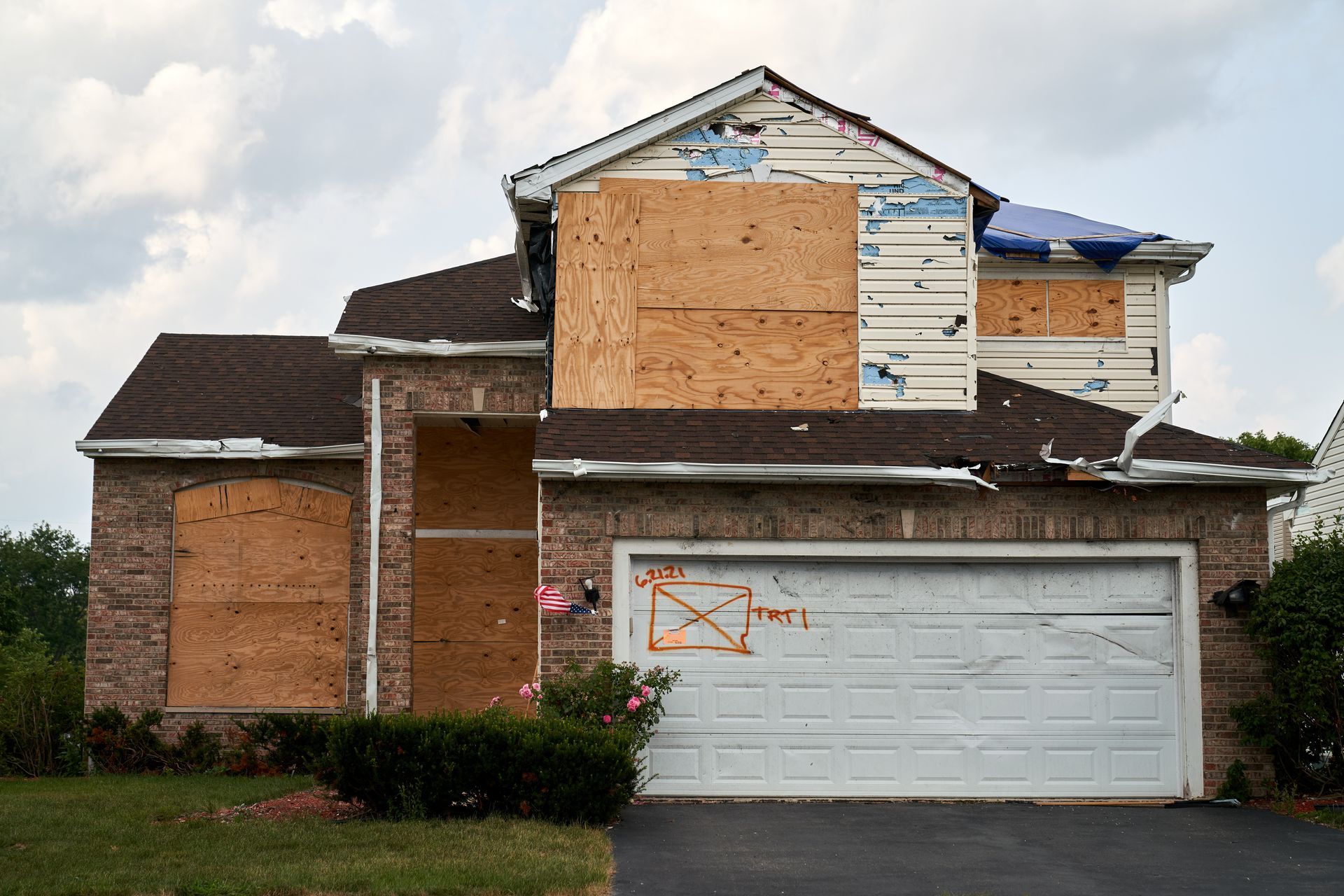 Partially boarded-up two-story suburban house with a white garage door and brick exterior