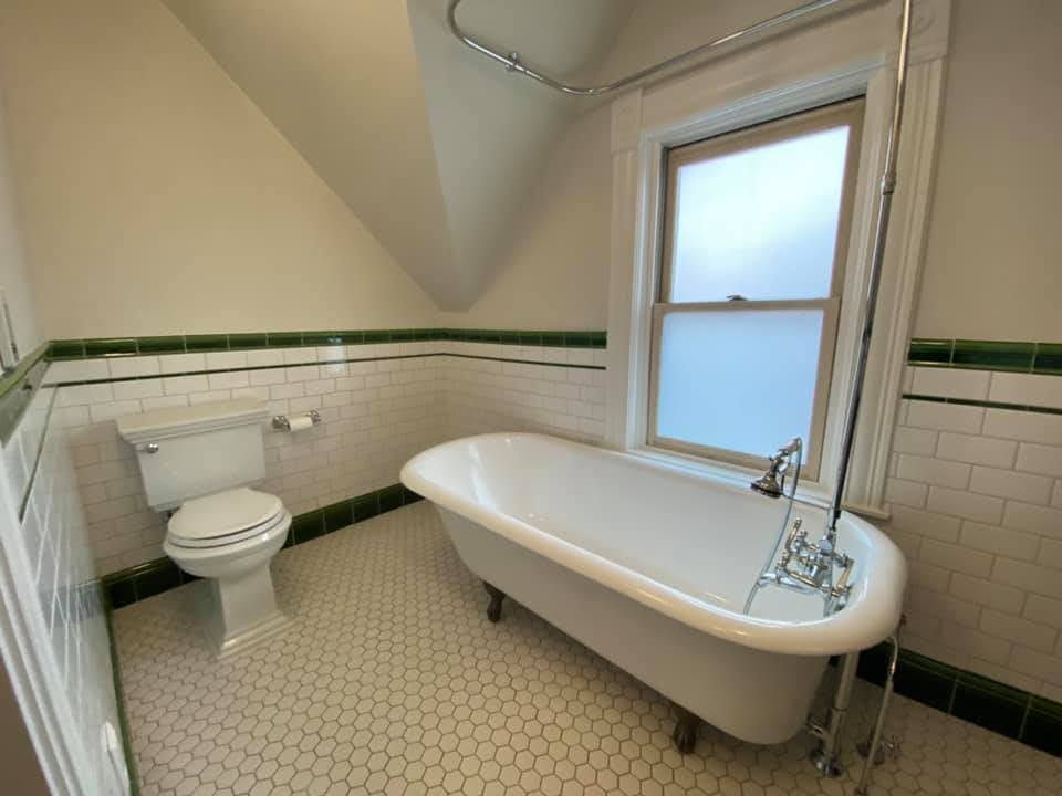 Small white bathroom with clawfoot tub, toilet, and green-accented tile walls under a sloped ceiling.