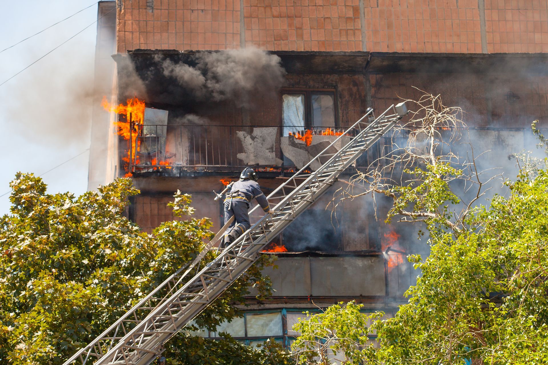 Firefighters climb a ladder toward a burning apartment building with smoke and flames pouring out.