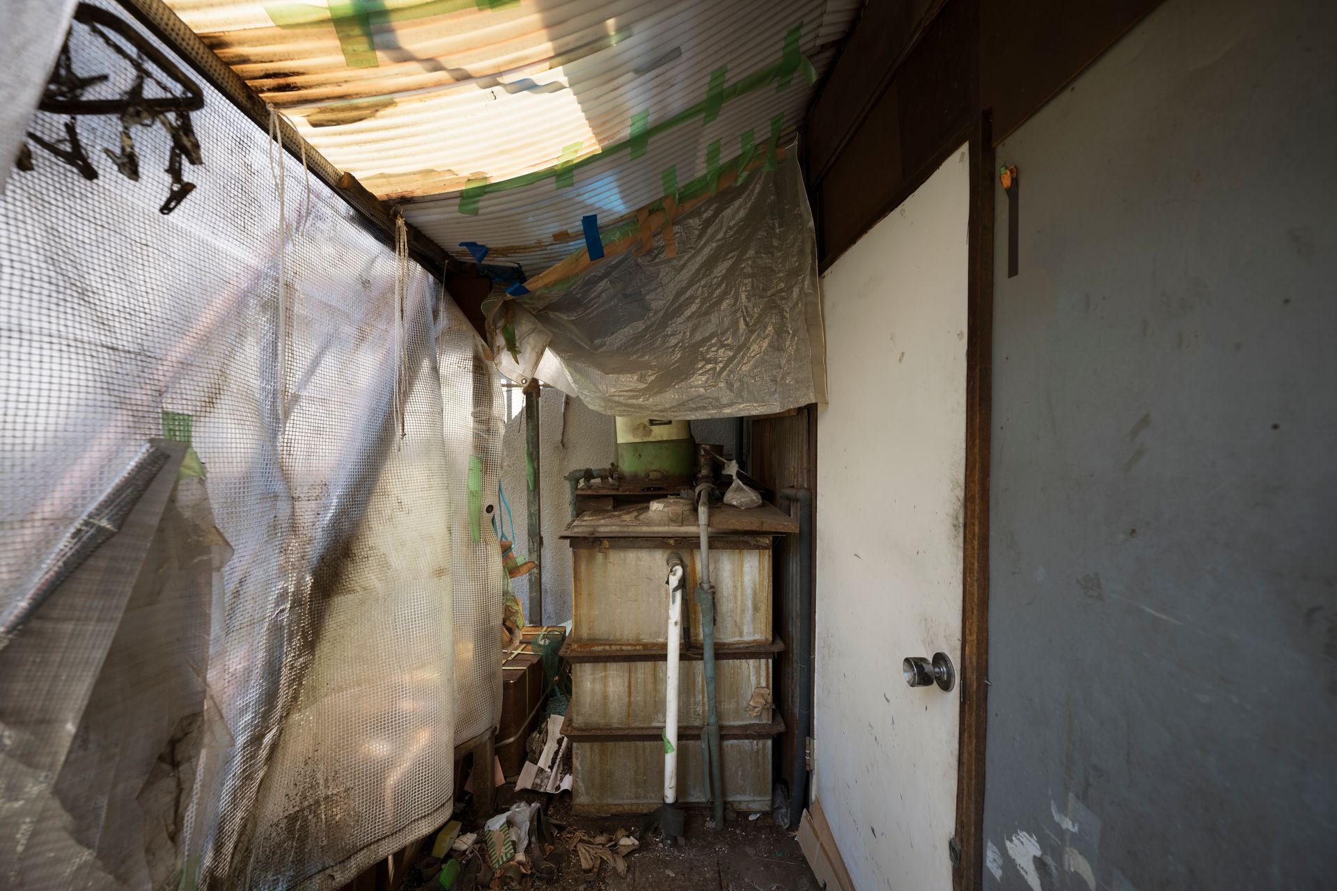 Narrow cluttered hallway with hanging plastic sheeting, a metal shelf, and a closed gray door
