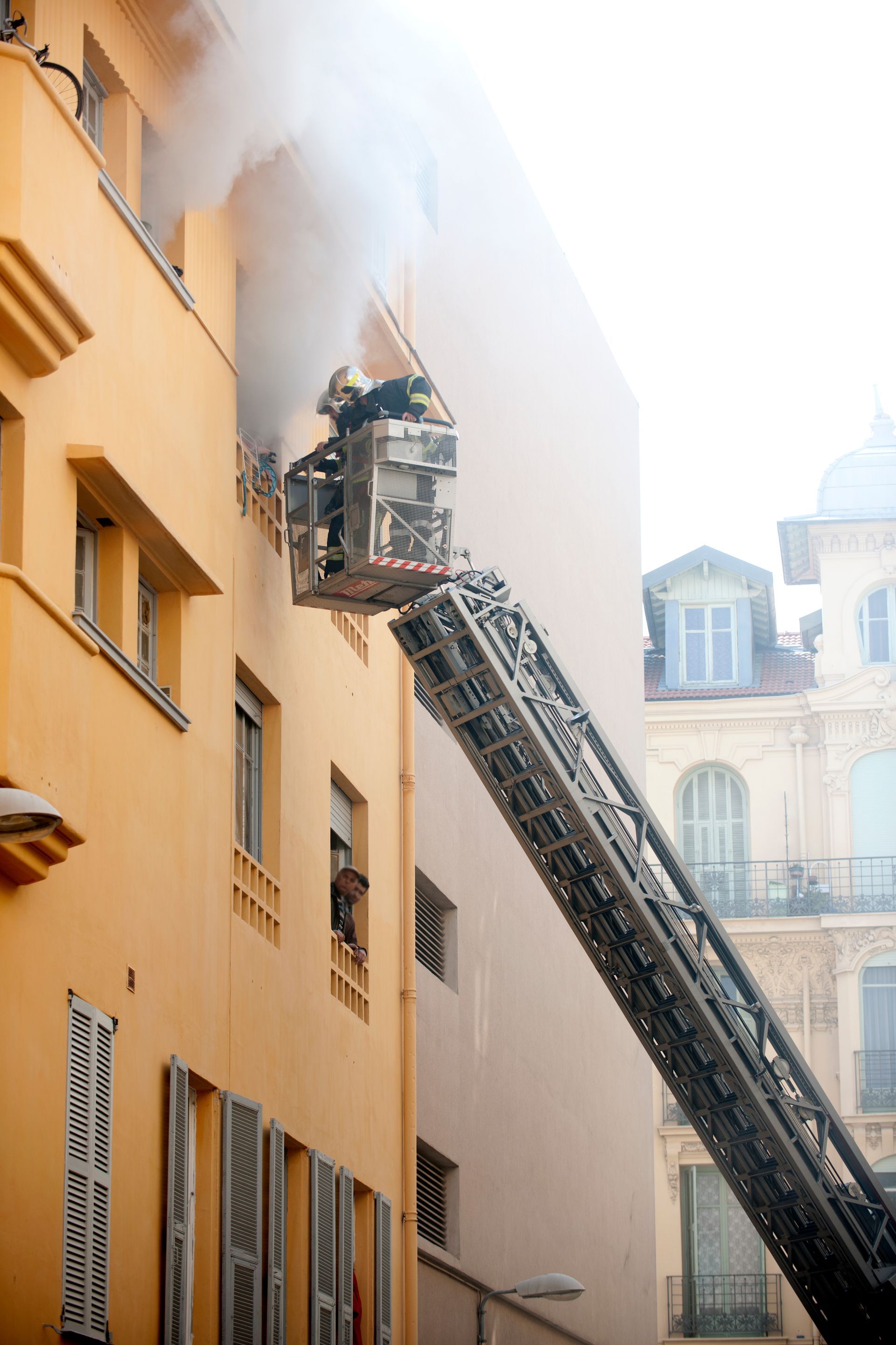 Firefighters on an extended ladder spraying water at a smoking upper-floor window on a narrow street