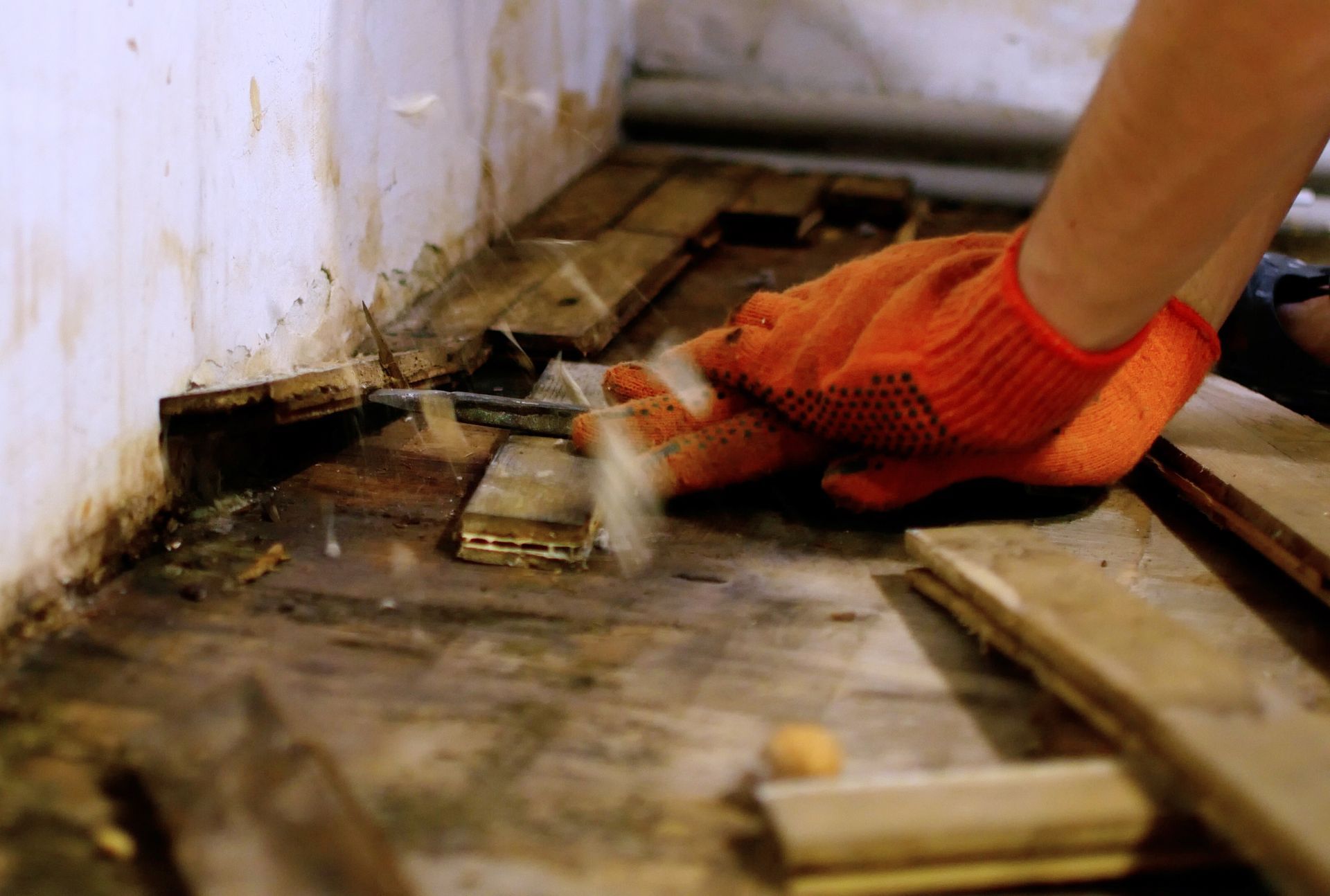 Gloved hand scraping debris from a dirty floor against a wall with exposed wood framing