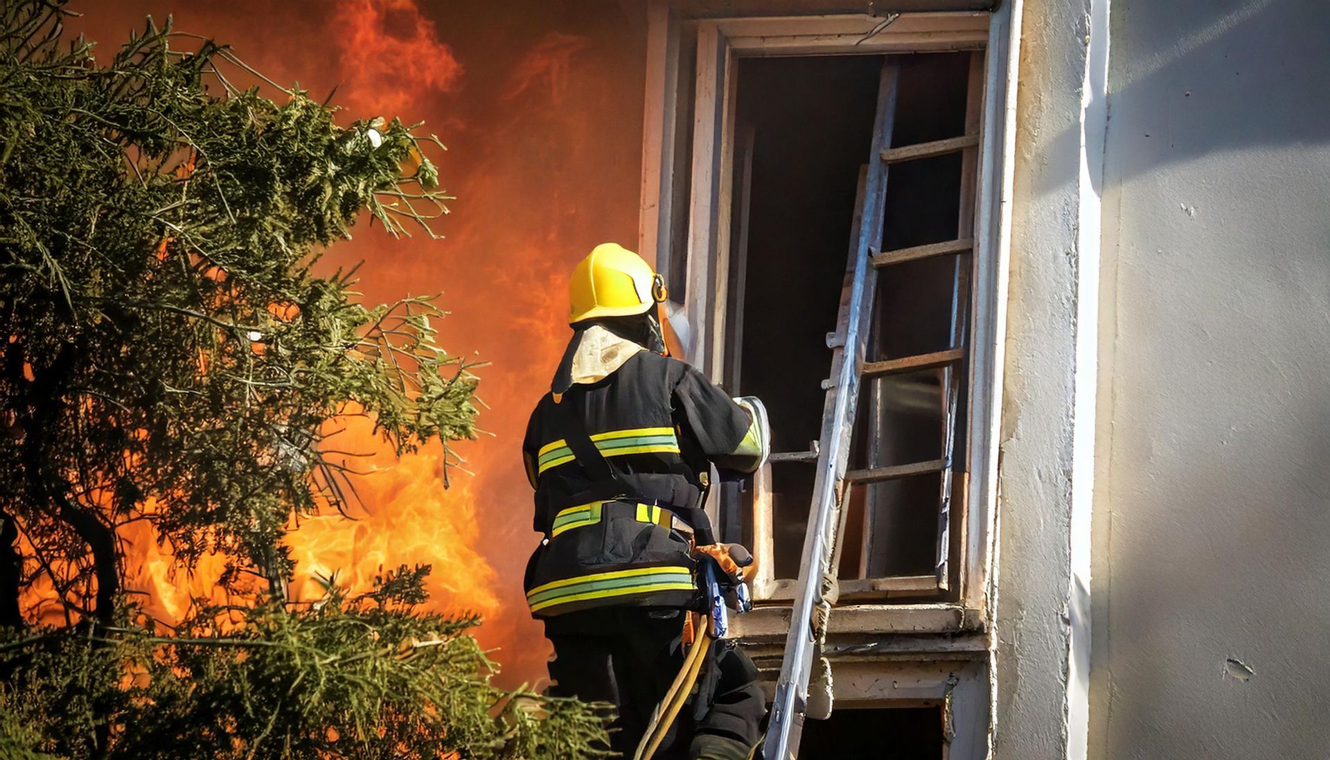 Firefighter battling flames beside a house, standing near a ladder and window.