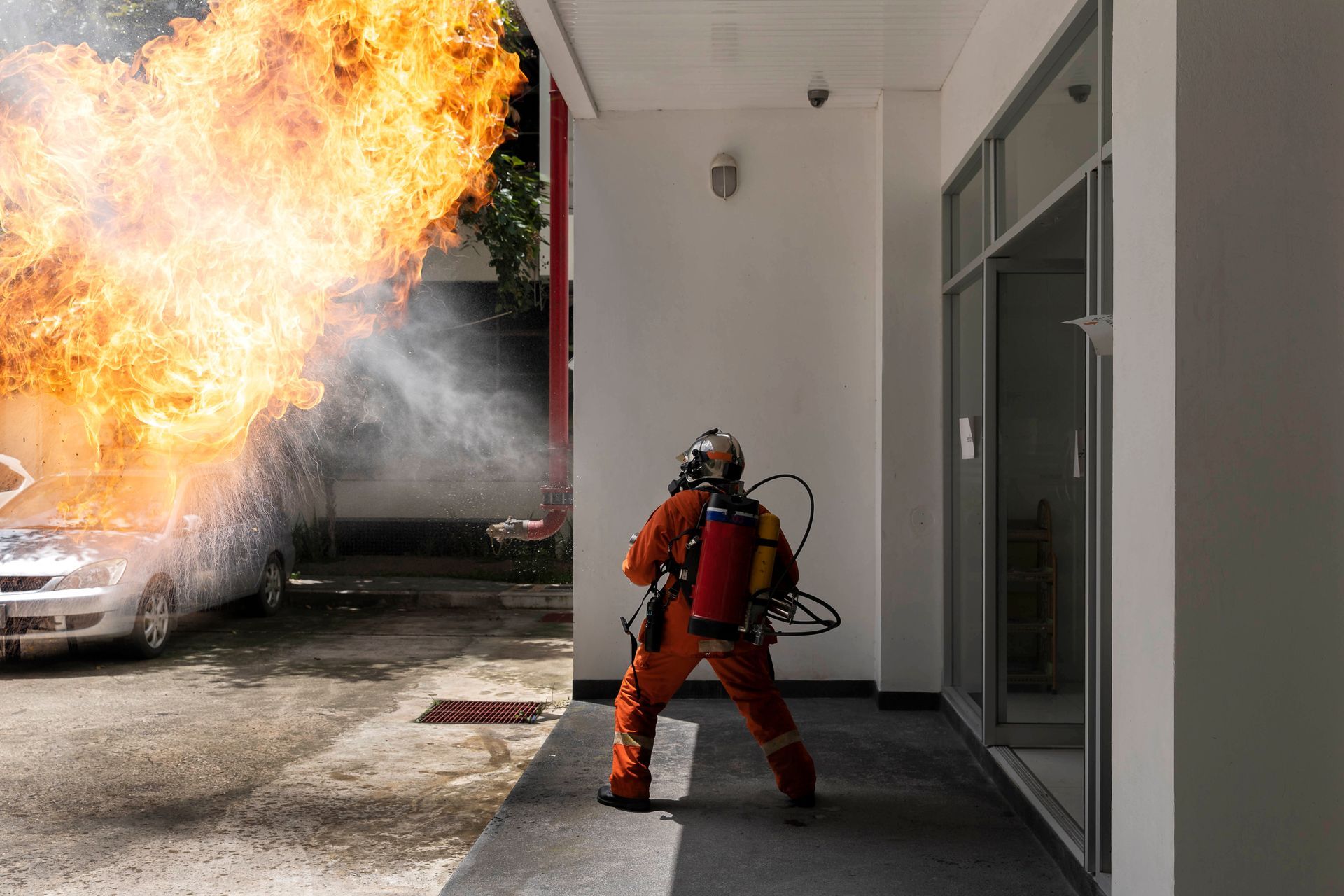 Firefighter facing a car fire with large flames outside a building entrance