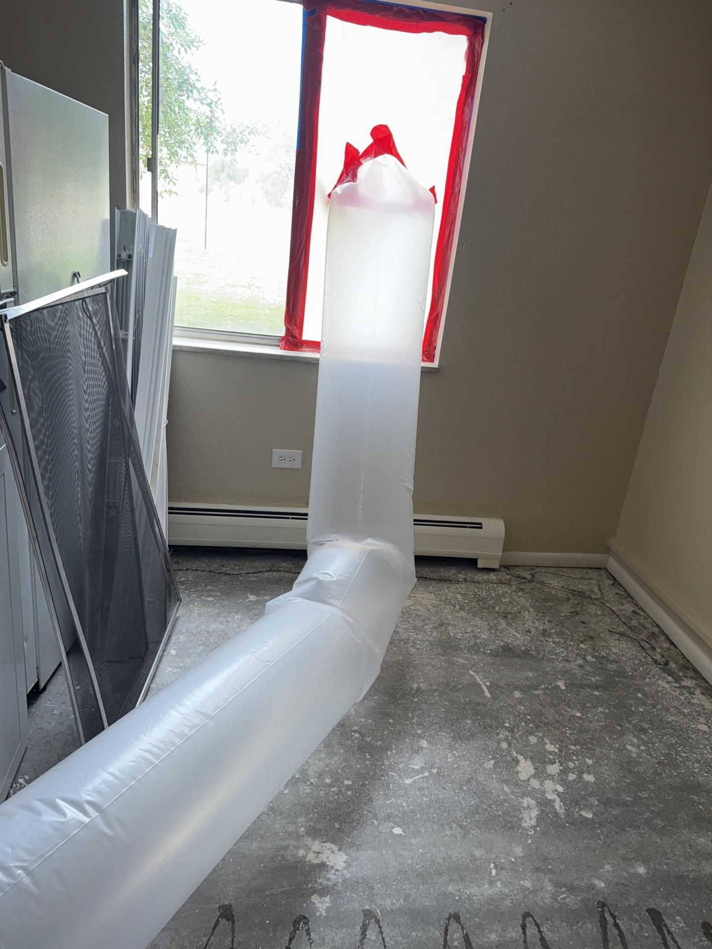 White flexible ducting runs across a dusty room to a window, with red tape sealing the opening.