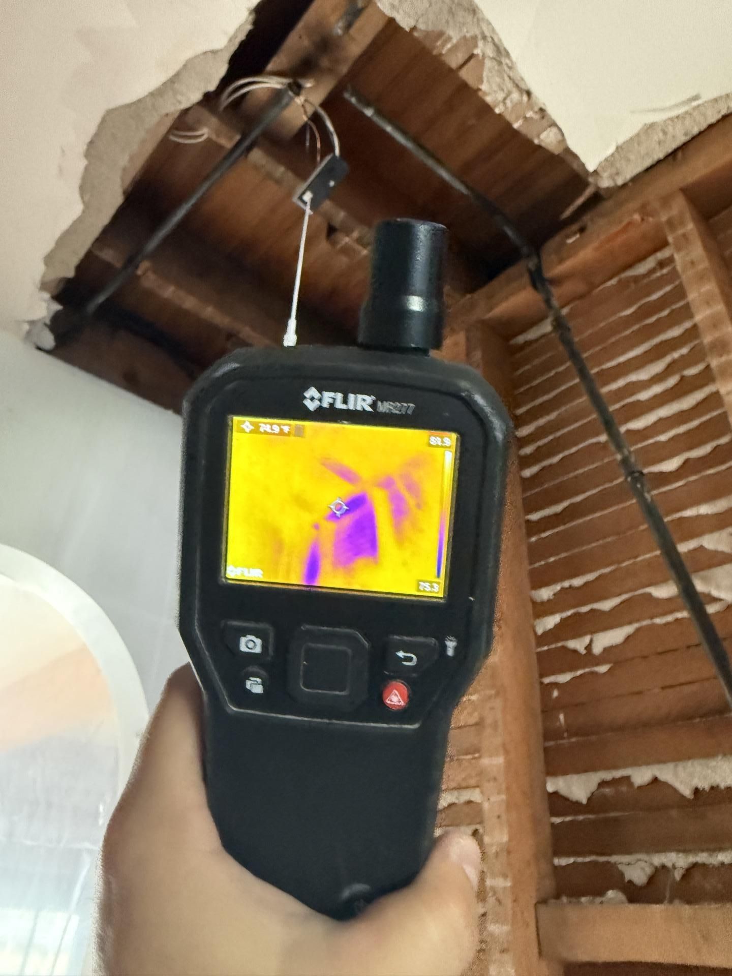 Handheld thermal camera pointed at a ceiling, showing heat patterns near exposed wooden beams and wiring