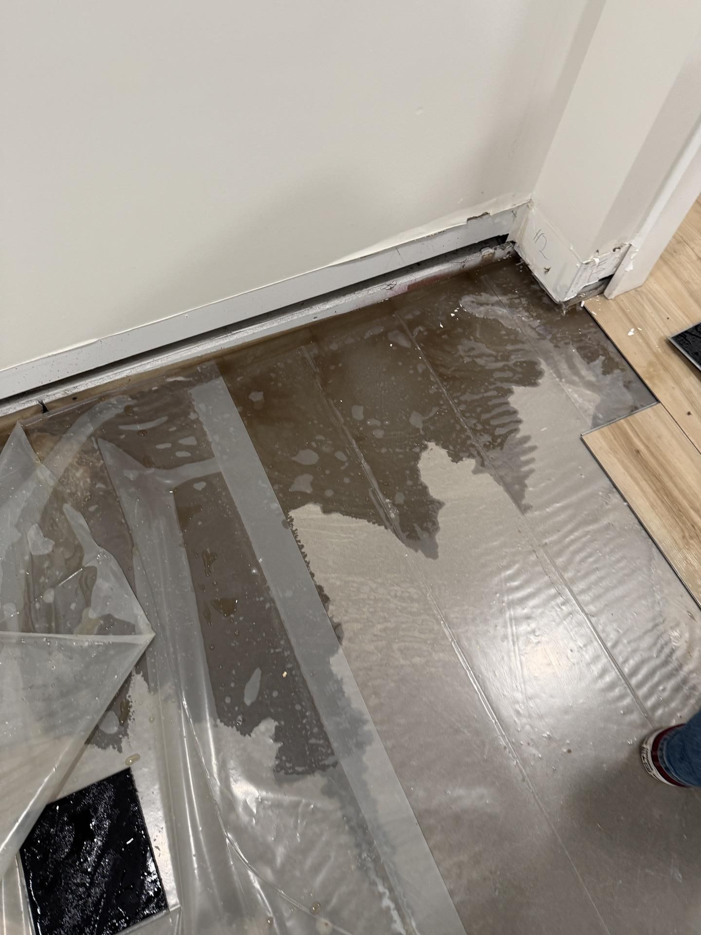 Water-damaged floor near a wall and doorway, with plastic sheeting and exposed underlayment.