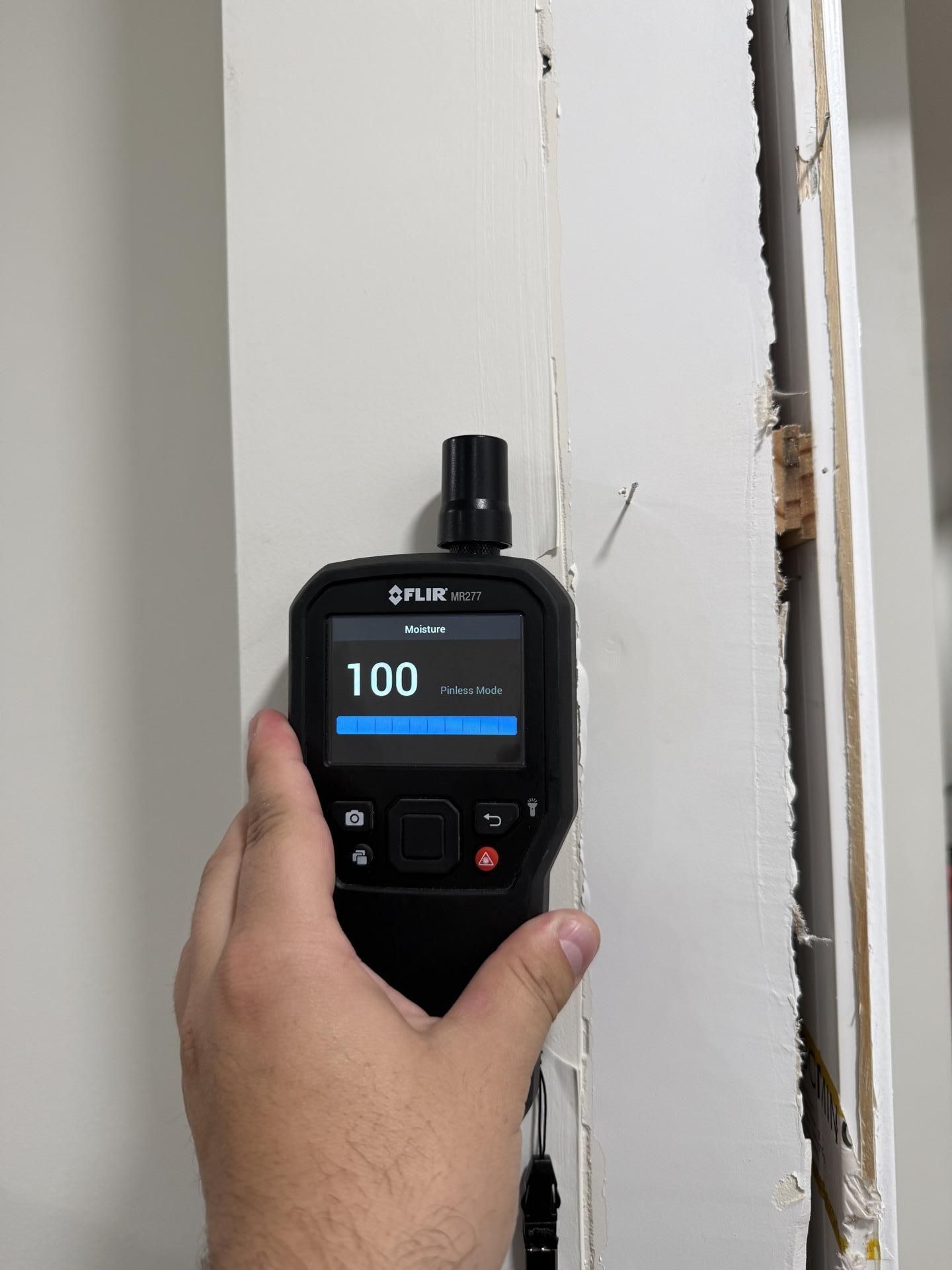 Handheld moisture meter reading 100 on a white wall near visible water damage and peeling paint